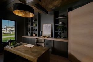 Dark home office with built-in shelving, wooden desk, gold pendant light, and large window overlooking a green landscape.