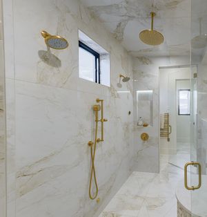 Luxury marble shower with gold fixtures, dual rain heads, handheld sprayer, and a small window for natural light.