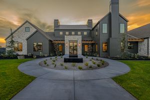 Modern farmhouse exterior at dusk with circular driveway, stone accents, and lush green lawn under dramatic sunset sky.