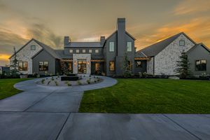 Modern farmhouse with stone and dark siding, circular driveway, lush lawn, and dramatic sunset sky.