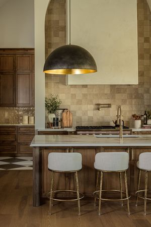 Elegant kitchen with dome pendant light, wood island, white barstools, brass accents, and neutral tile backsplash.