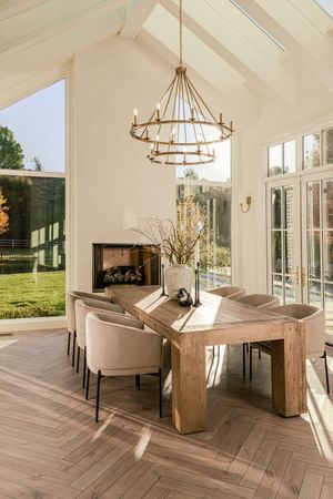 Sunlit dining room with vaulted ceiling, rustic wood table, upholstered chairs, tiered chandelier, fireplace and herringbone wood floors.