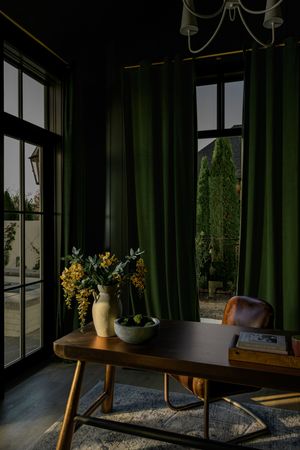 Moody home office with deep green velvet curtains, a wooden desk, leather chair, yellow flowers in a cream vase and sunlit garden windows.