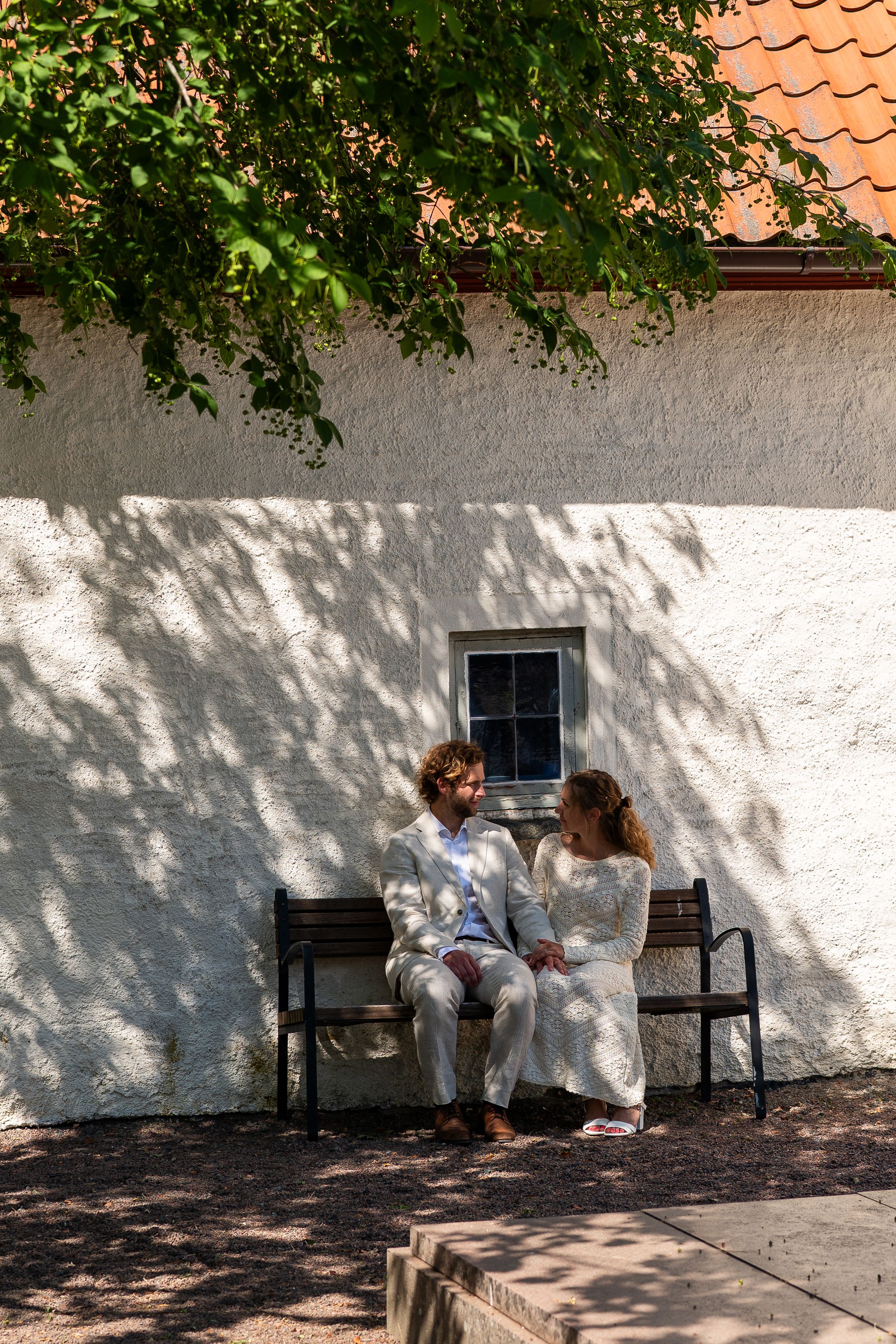 A couple in wedding attire sits on a bench against a white wall, sharing a tender moment in dappled sunlight.