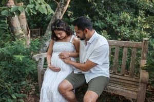 Pregnant couple sharing a tender moment on a wooden bench surrounded by lush greenery in an outdoor maternity photoshoot.