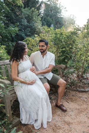 Pregnant woman in white dress sits with bearded man on garden bench, surrounded by lush green foliage.