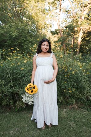 Pregnant woman in white maxi dress holding sunflower bouquet, posing outdoors in lush green garden setting.