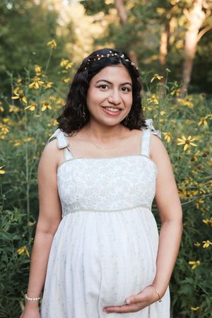 Smiling pregnant woman in white floral dress with hair crown poses among yellow wildflowers outdoors.