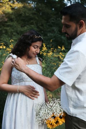 Pregnant woman in white dress holds sunflowers while man tenderly touches her belly outdoors.