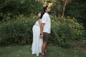 Pregnant couple standing back-to-back in a garden, woman in white dress, man in white shirt, surrounded by yellow flowers.