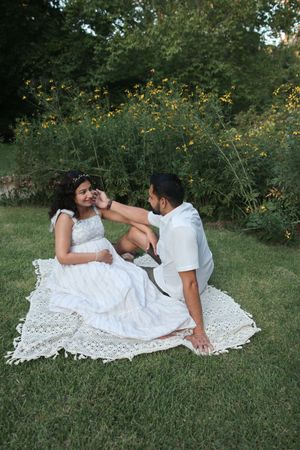 Couple sits on lace blanket in garden, man gently touches pregnant womans face affectionately.