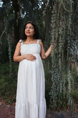 Pregnant woman in white floral maxi dress poses near weeping tree, gazing upward gracefully.