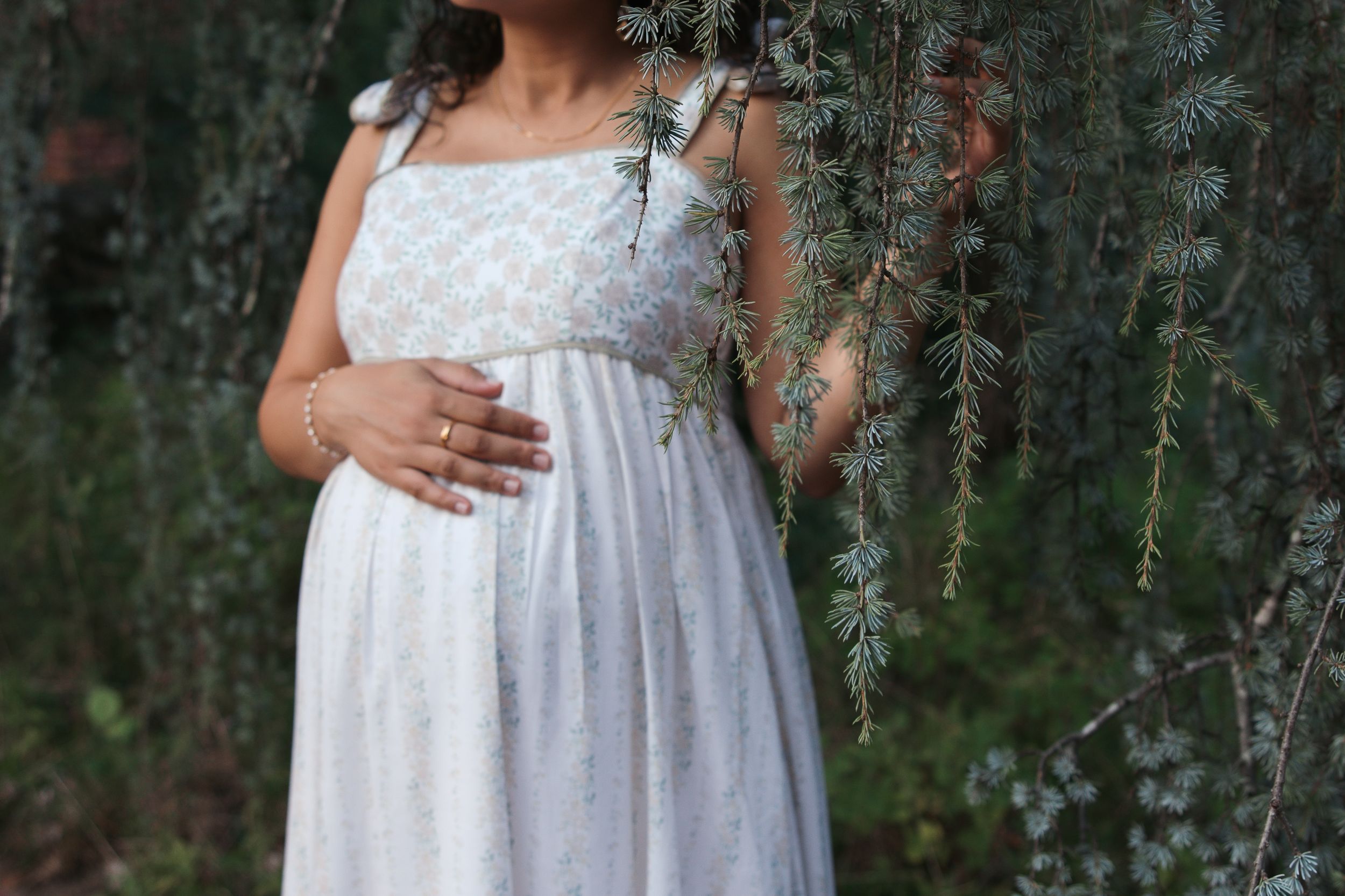 Pregnant woman in white floral dress cradling baby bump outdoors among lush green pine tree branches.