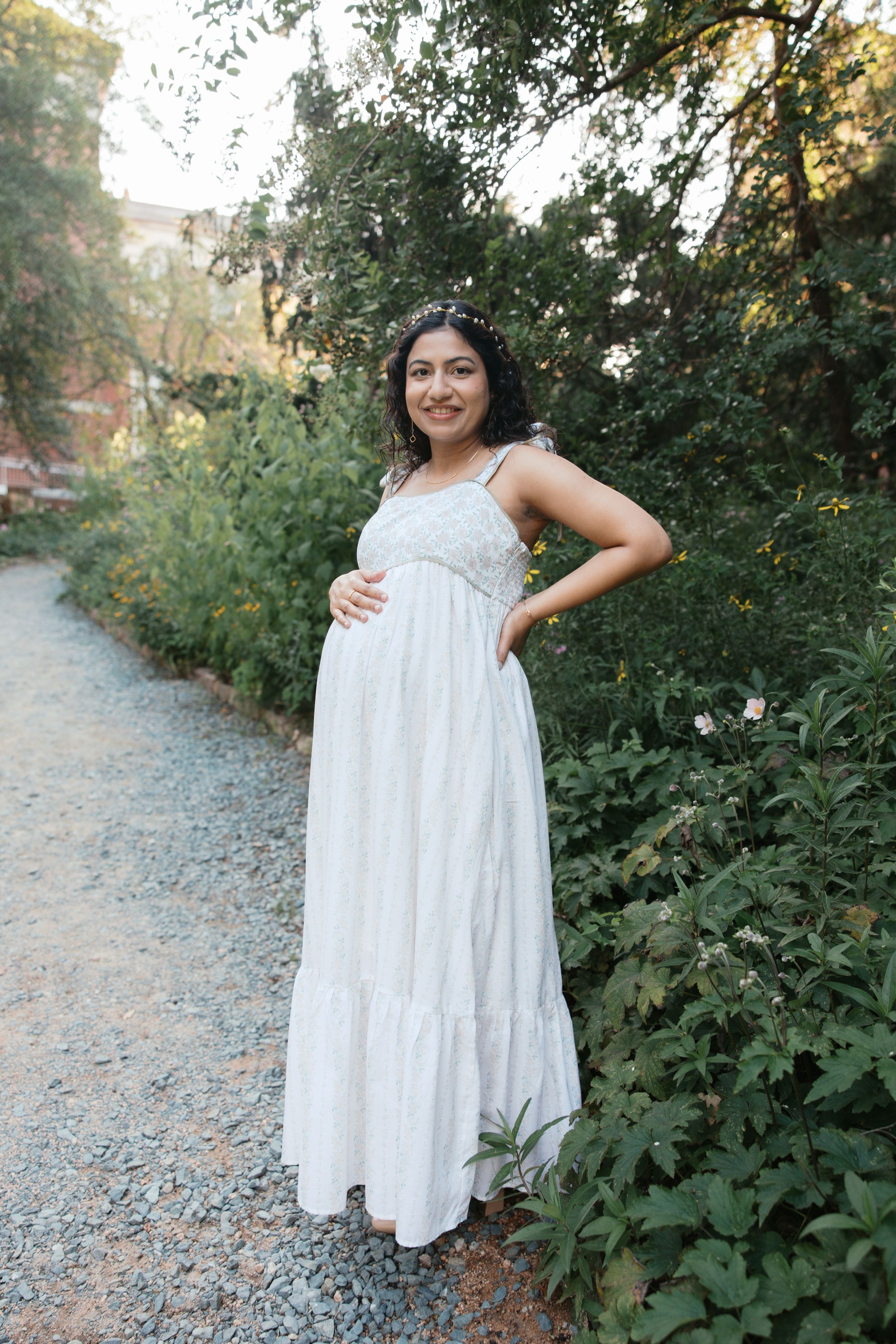 Pregnant woman in white maxi dress posing outdoors among lush green plants on a gravel path.