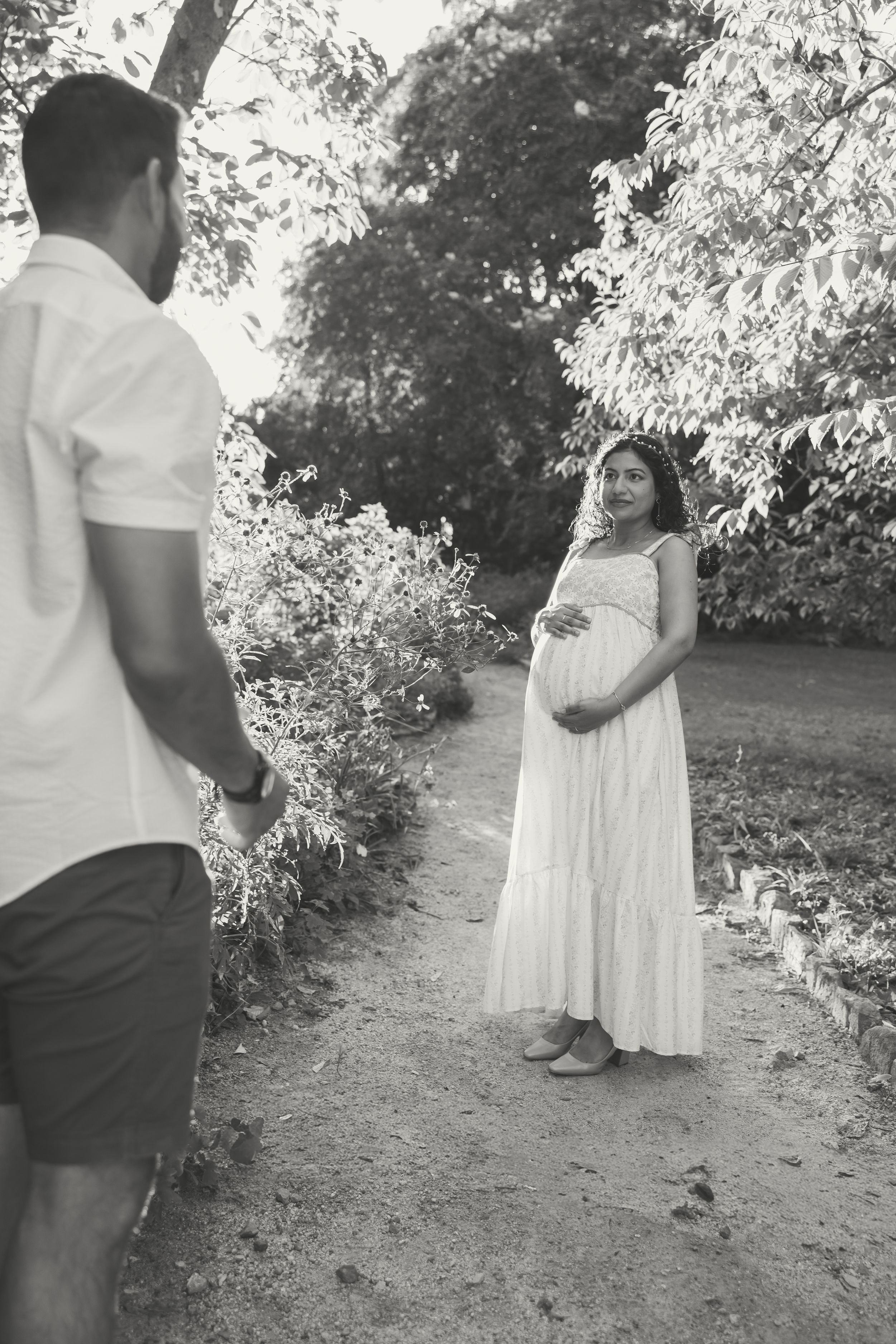 Black and white photo of a pregnant woman in a white dress holding her belly, facing a man on a garden path.
