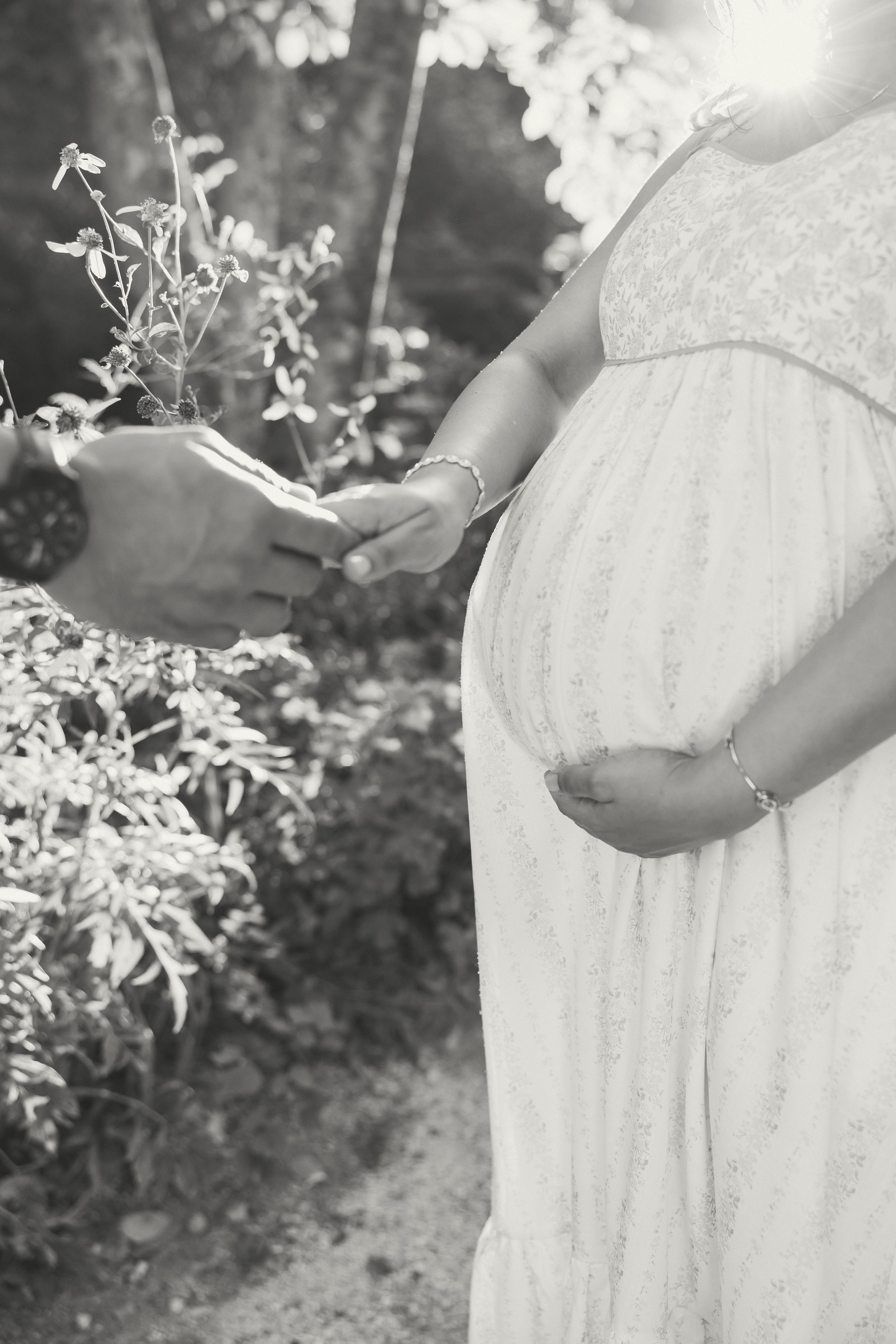 Black and white photo of a pregnant woman in a white dress holding her baby bump outdoors.