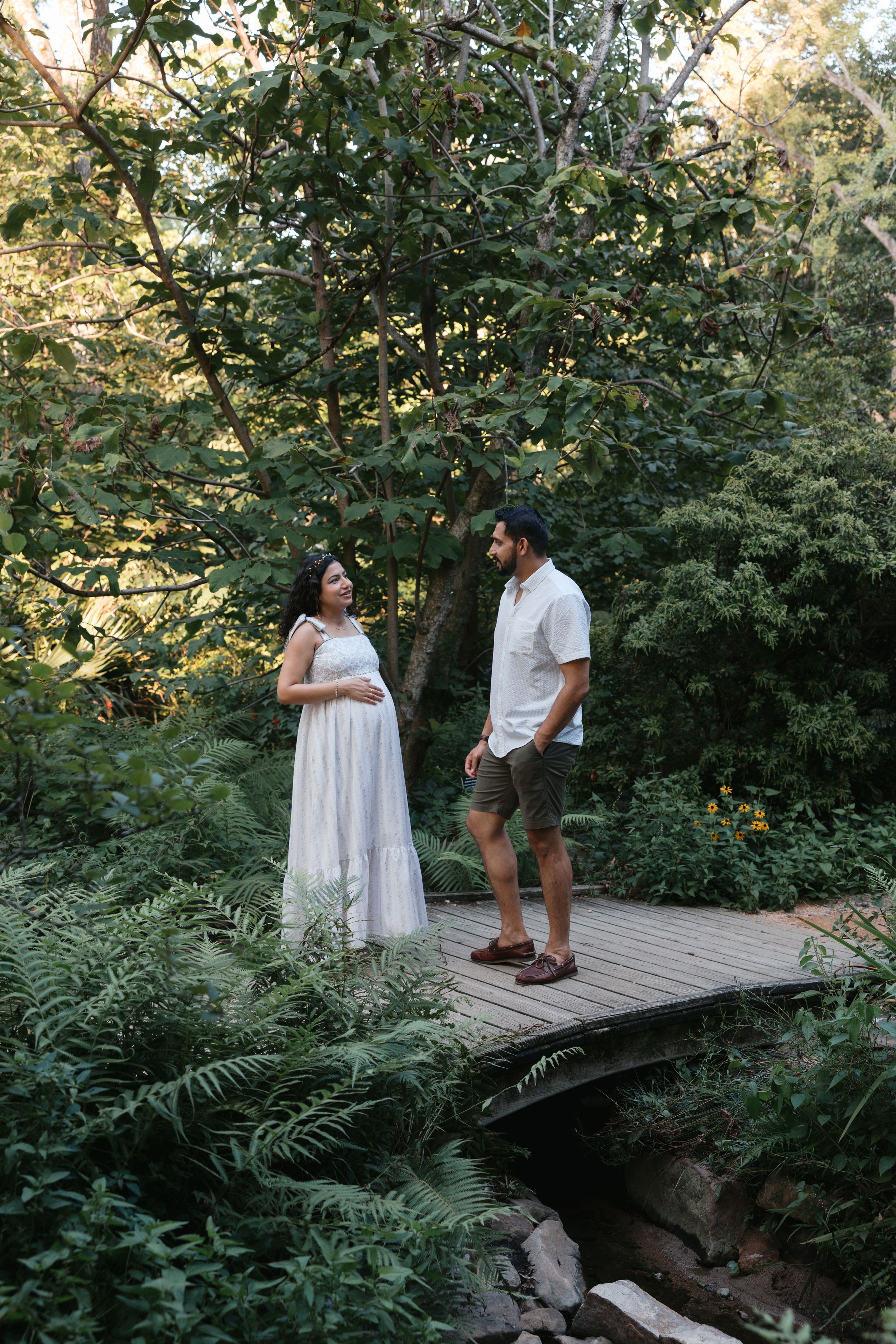 Pregnant woman in white dress with partner on wooden bridge surrounded by lush green forest trees and ferns.