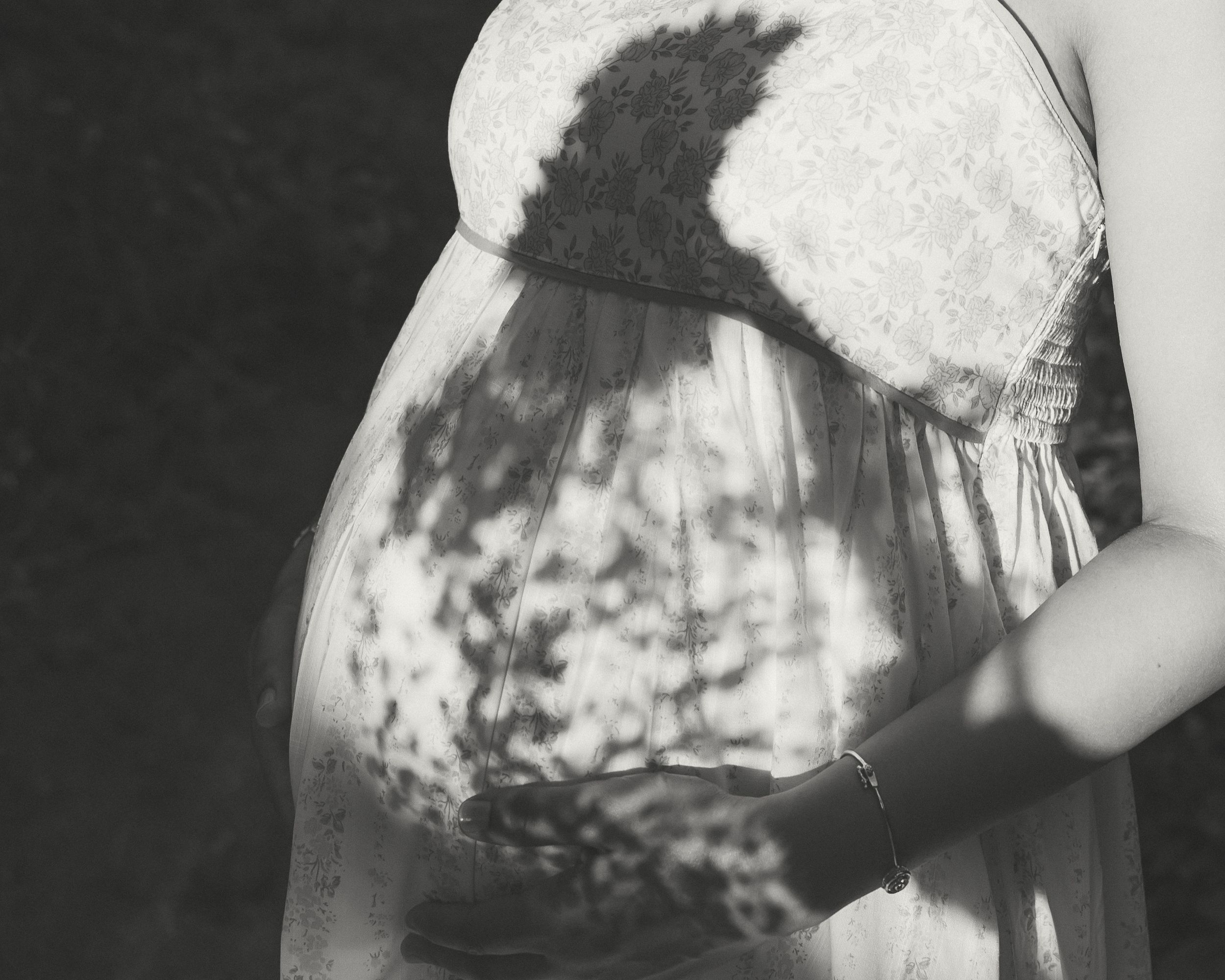 Black and white maternity photo of pregnant woman in lace dress with dappled sunlight shadows on her baby bump.