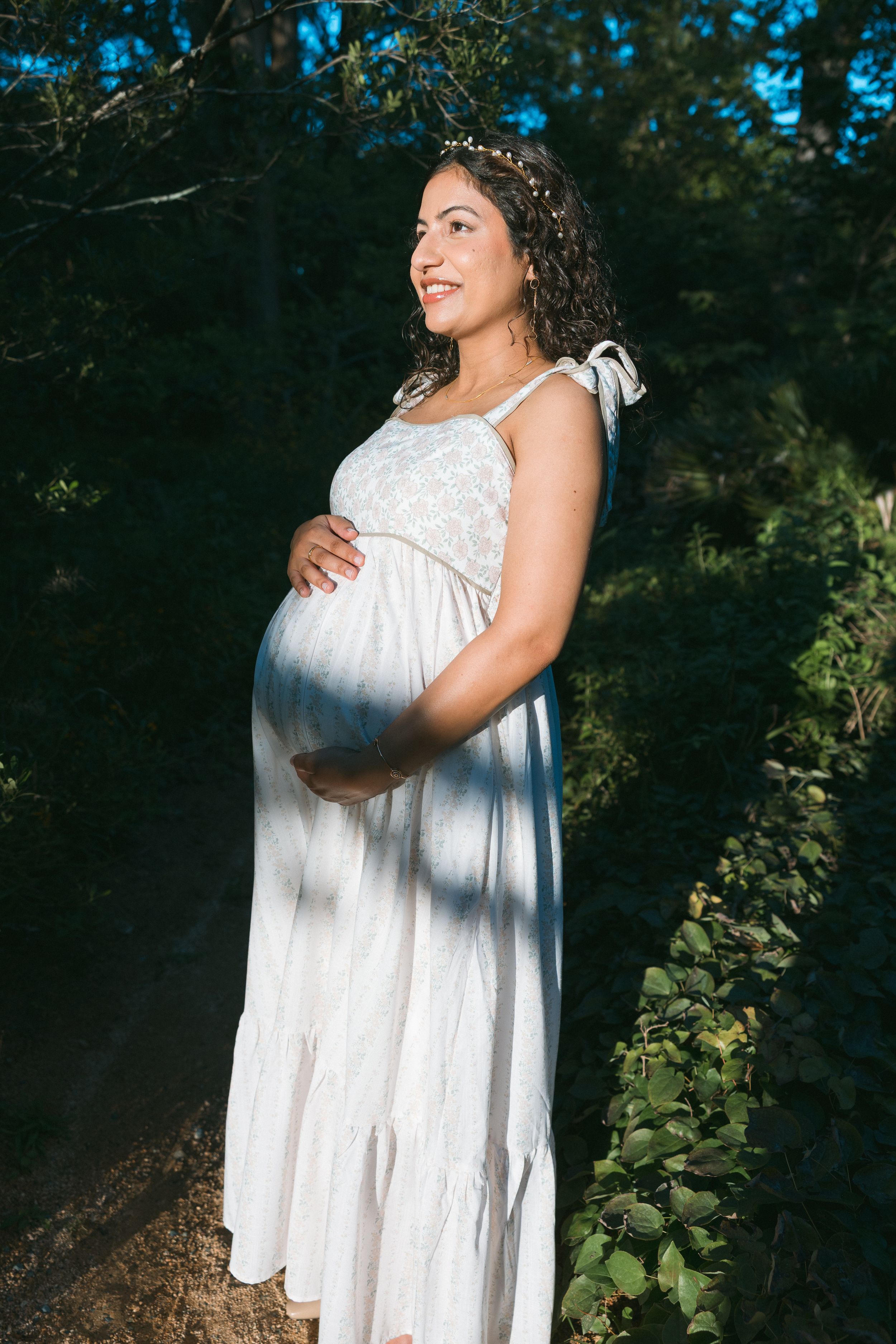 Smiling pregnant woman in white dress holding belly, wearing floral crown, standing outdoors in sunlit green forest.