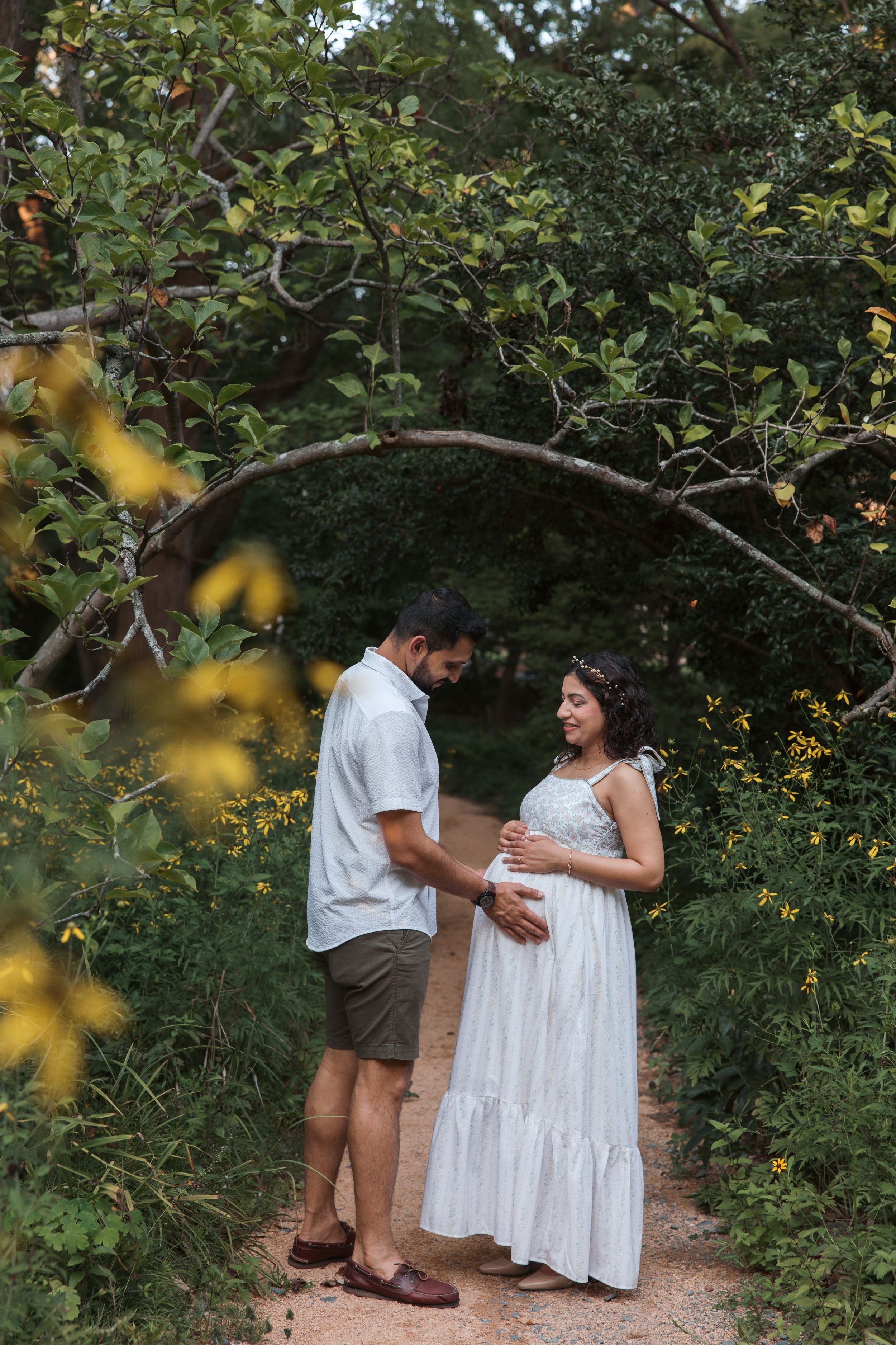 Couple sharing a tender moment on a garden path, pregnant woman in white dress under a natural tree arch.