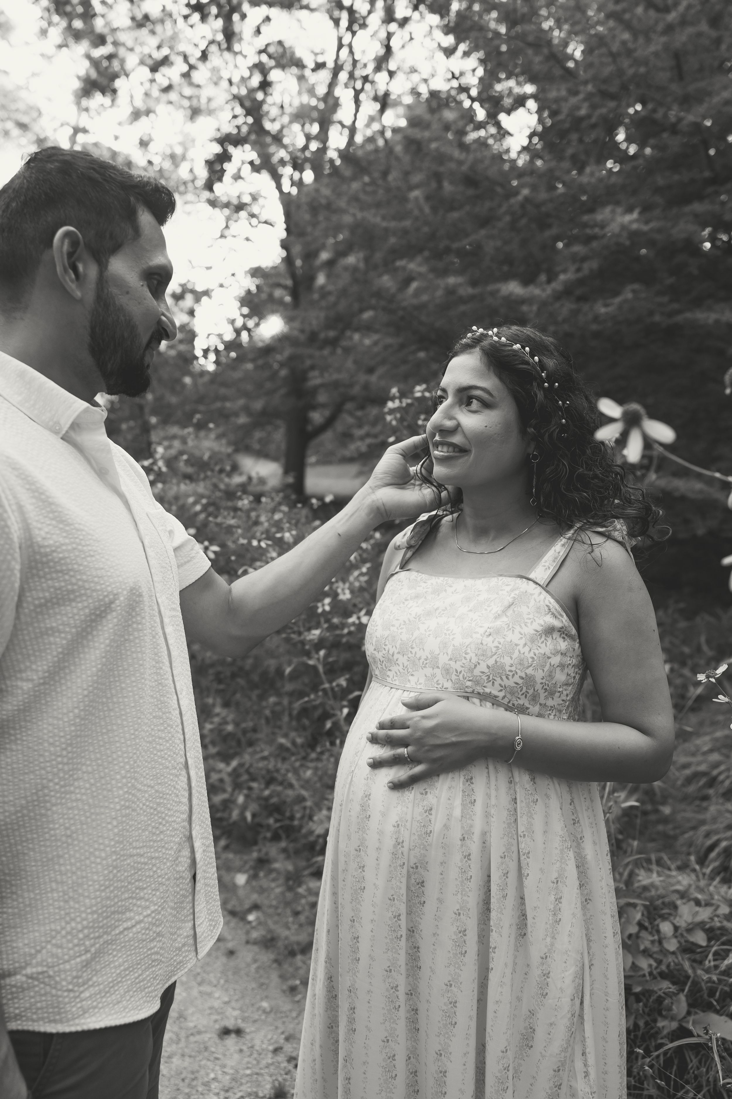 Black and white photo of a bearded man tenderly touching a smiling pregnant woman's face in a lush garden setting.