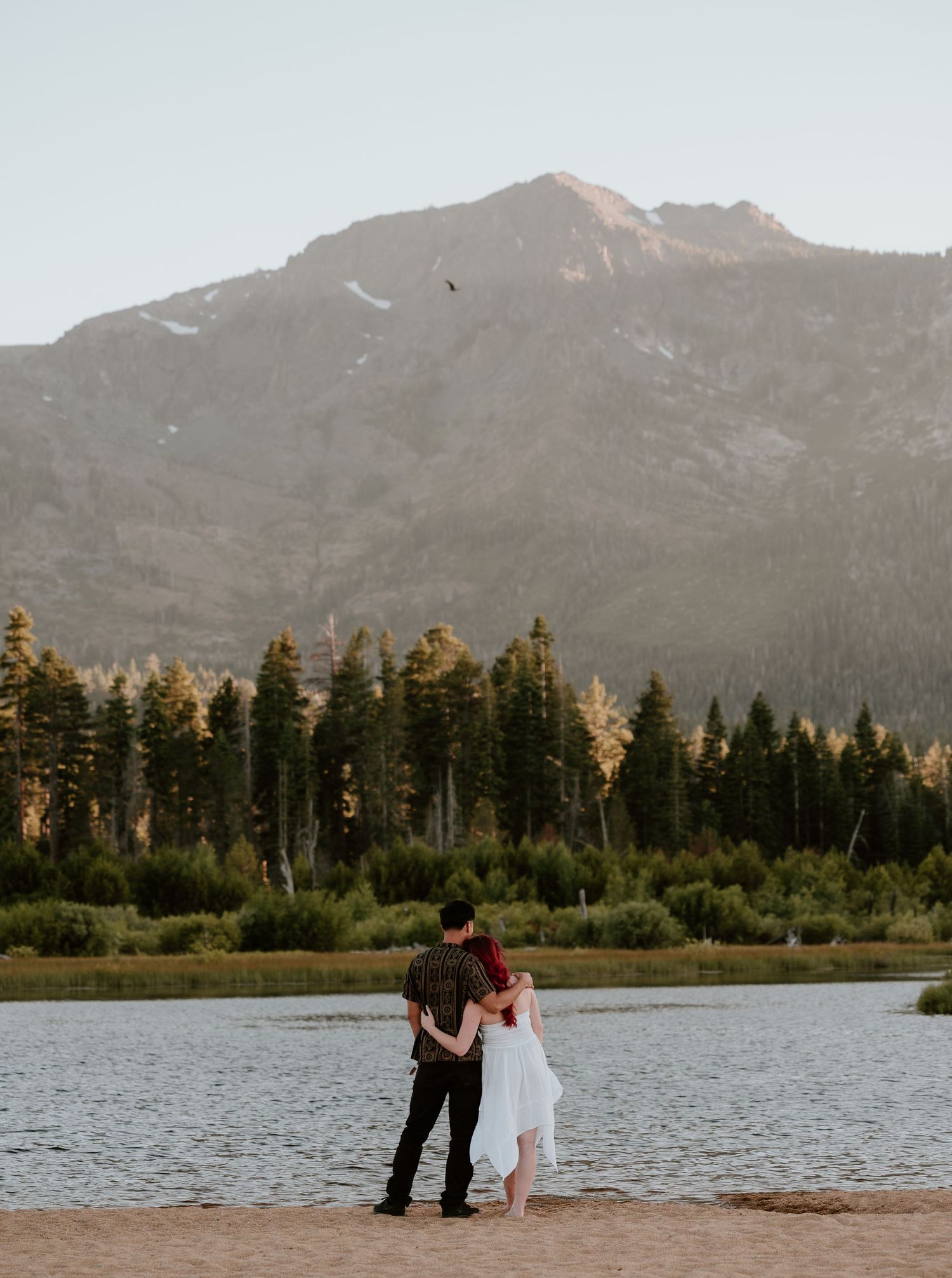 Couple embracing on sandy shore with mountain lake and pine forest backdrop at golden hour.