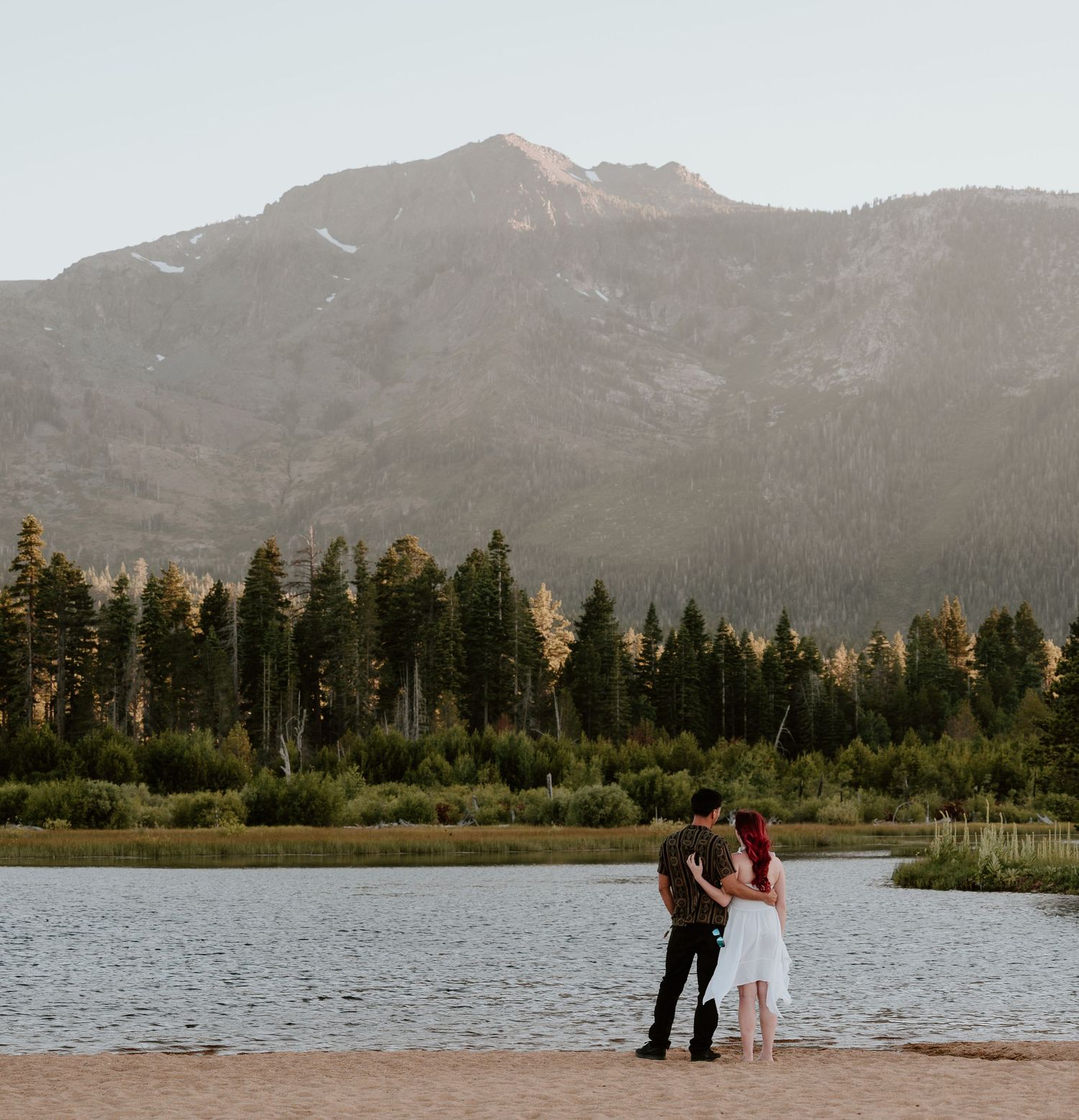 Couple embracing by a mountain lake with pine trees and rocky peaks in the background during golden hour.