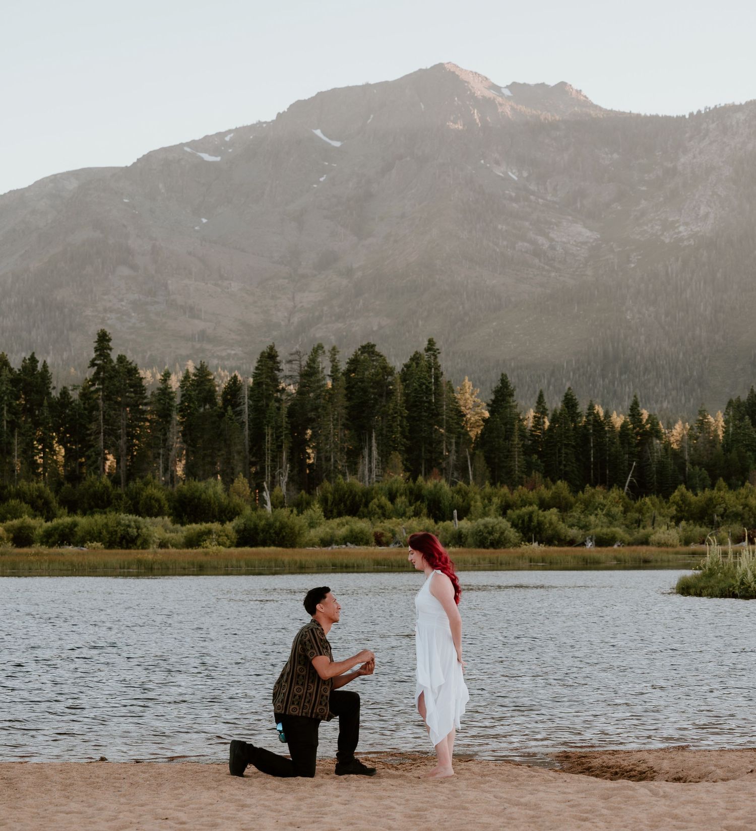 Man proposes on one knee to woman in white dress by mountain lake with pine trees backdrop.