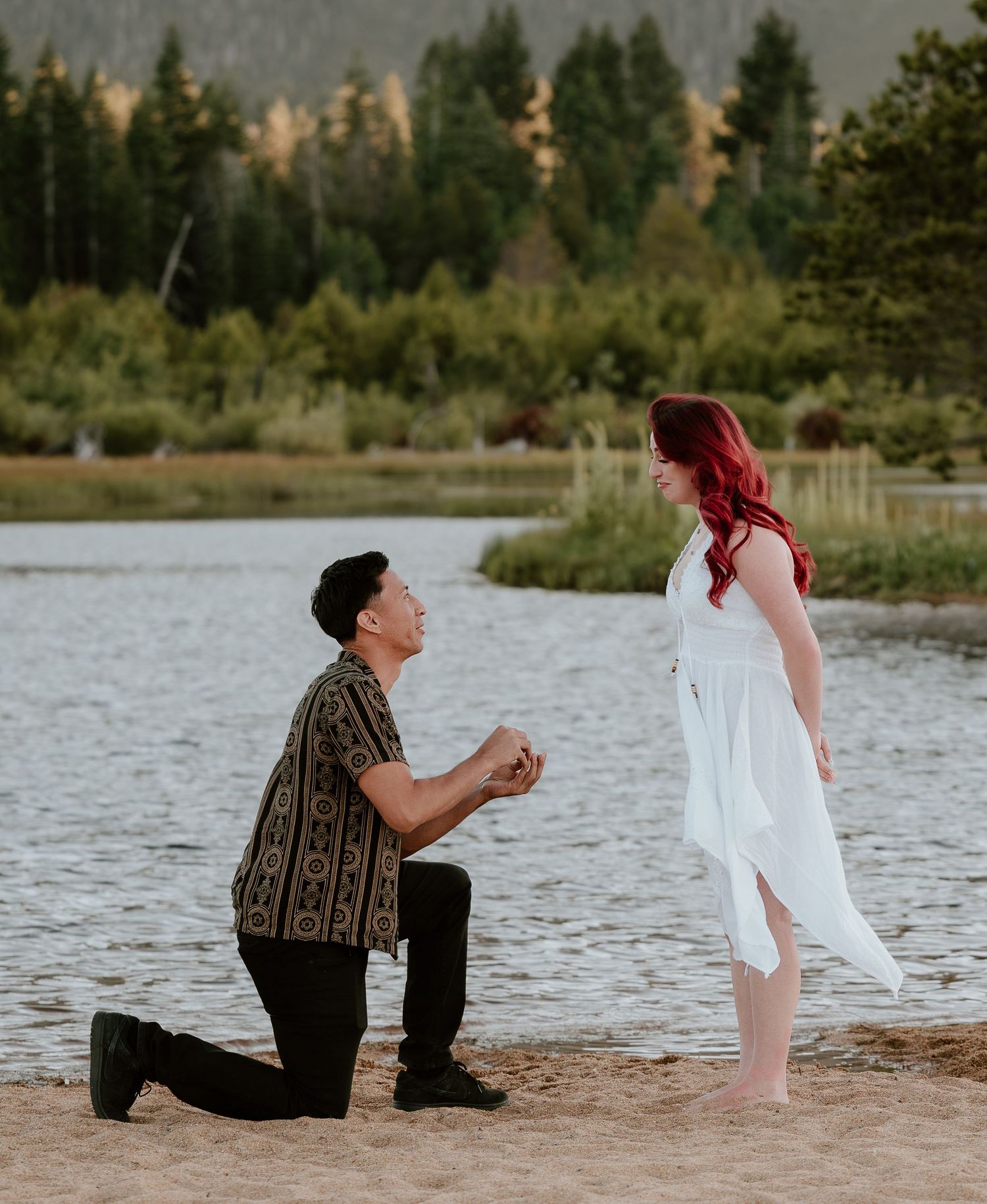 Man proposing on one knee to red-haired woman in white dress by a scenic mountain lake.