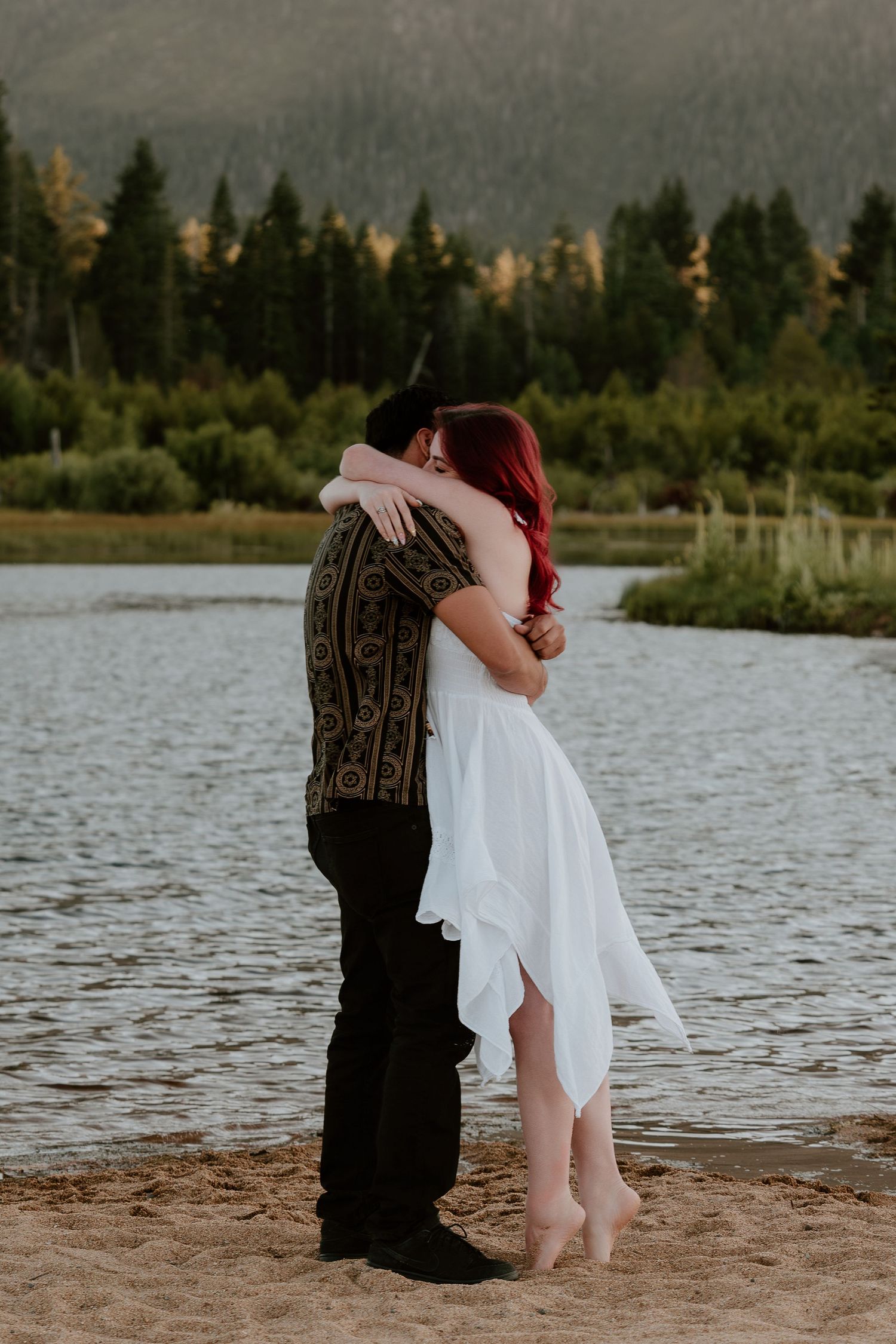 Couple embracing by a mountain lake, woman in white dress with red hair, surrounded by pine trees and sandy shore.