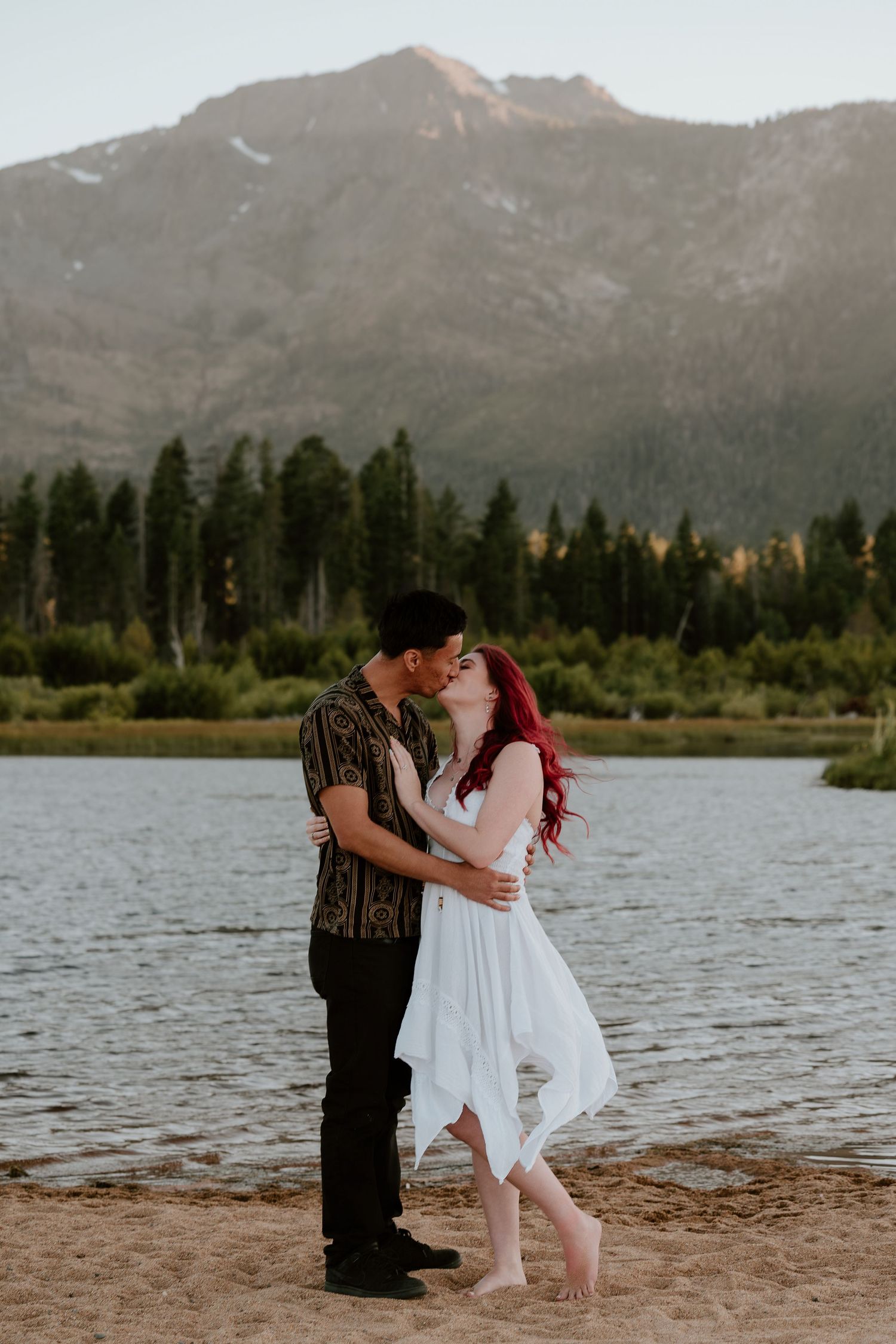 Couple kissing on sandy beach shore with mountain backdrop, woman in white dress with red hair.