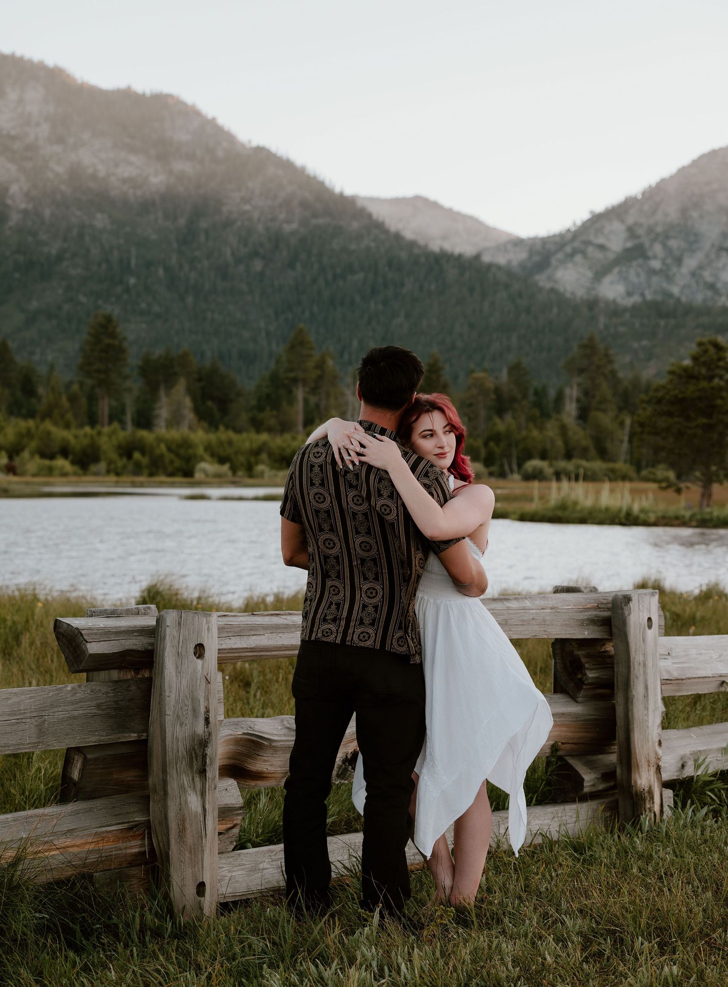 Couple embracing by a wooden fence with a mountain lake and pine trees in the background during golden hour.