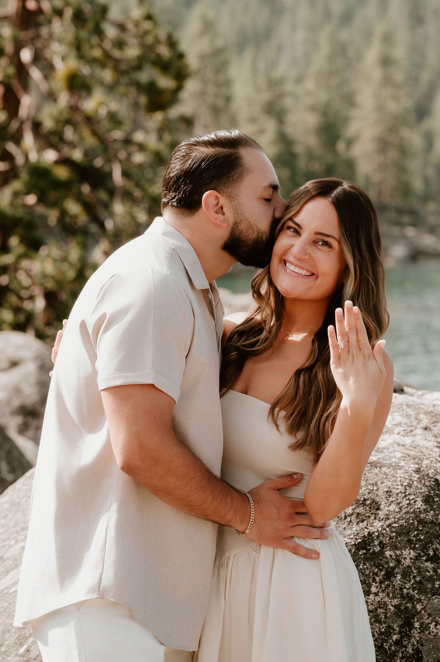 Couple in white outfits celebrating engagement outdoors, woman showing ring while man kisses her cheek near a lake.