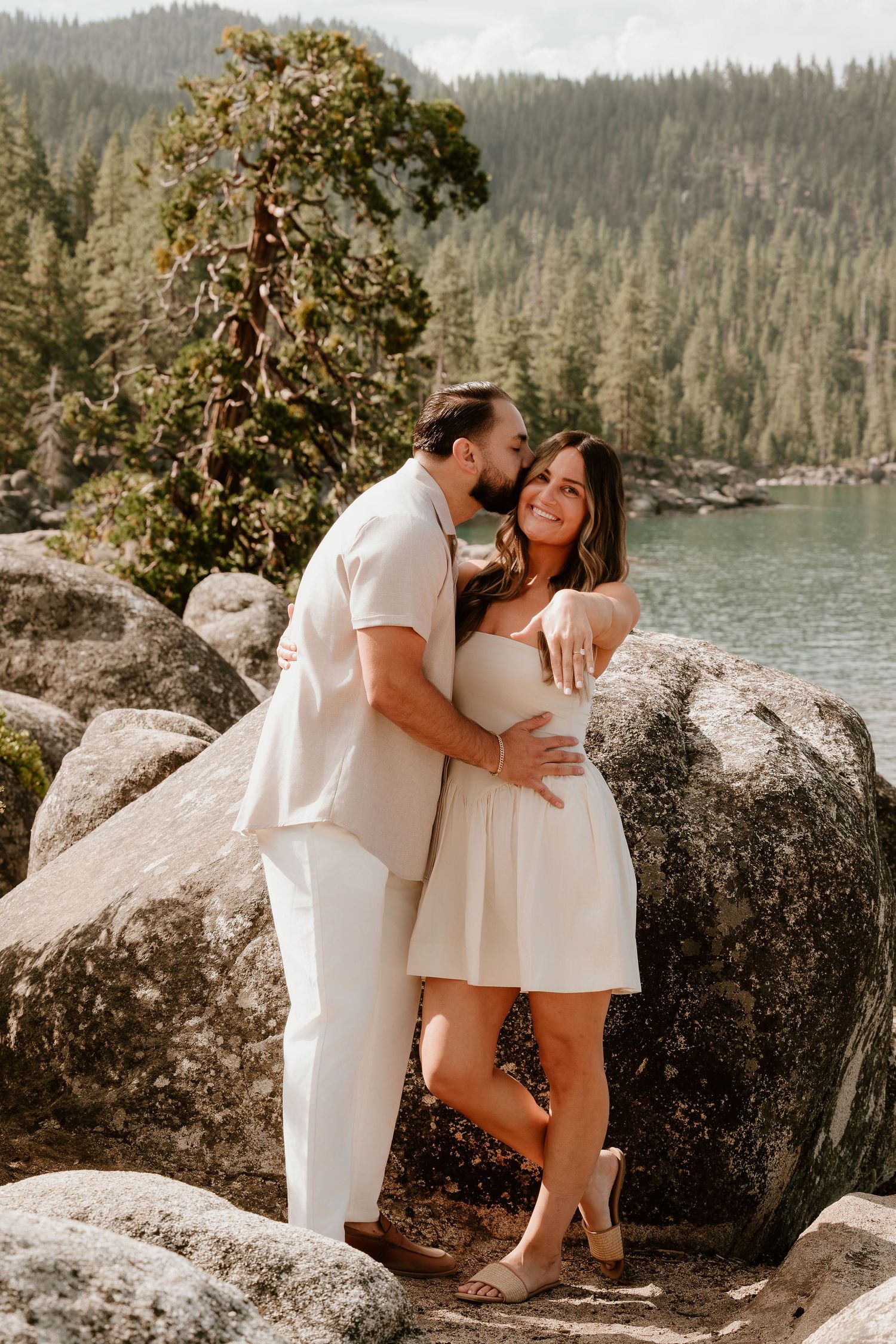 Couple in white outfits embracing on rocks by a mountain lake surrounded by pine trees.