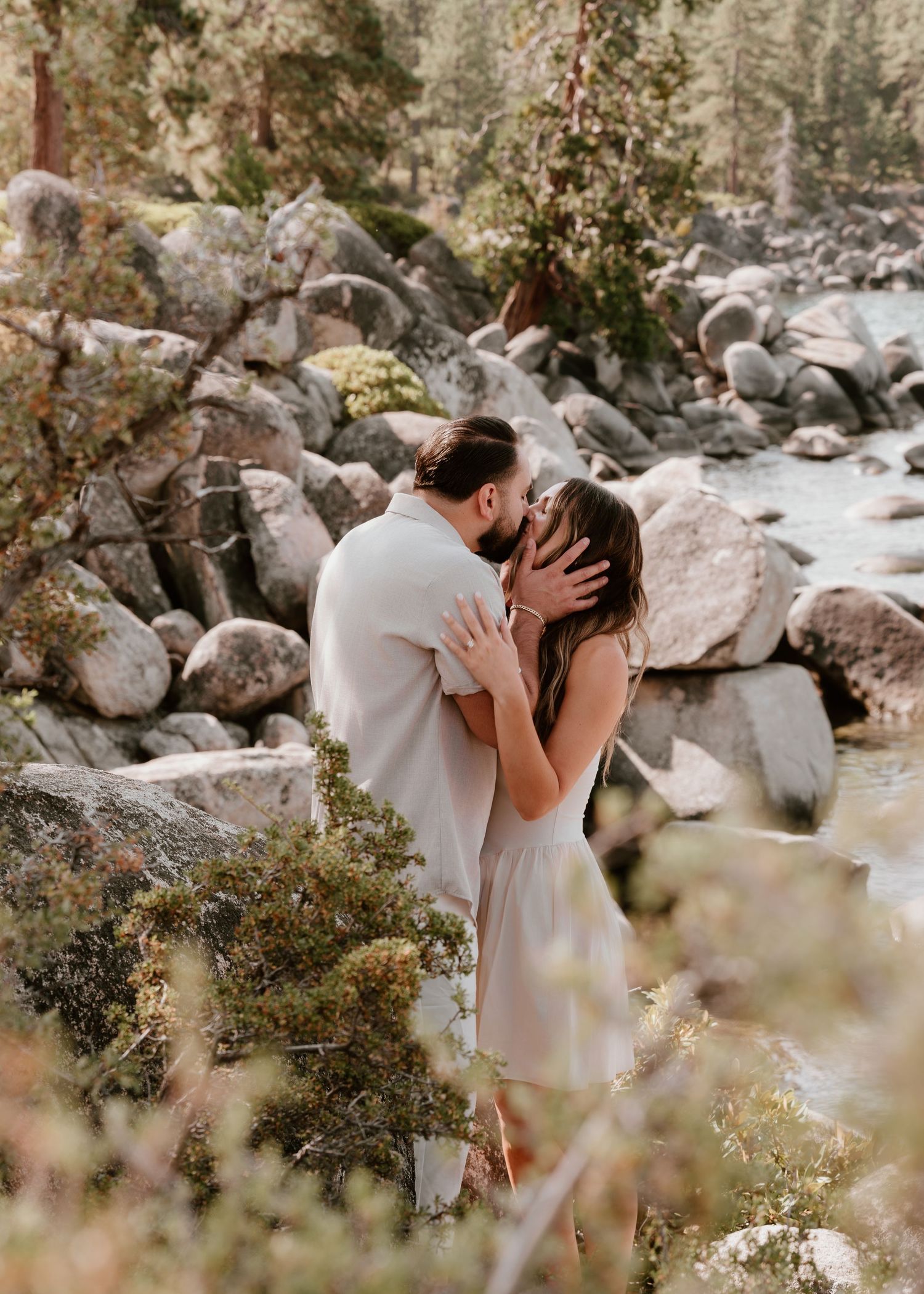 Couple sharing a romantic kiss among boulders and pine trees beside a mountain stream in a scenic outdoor setting.