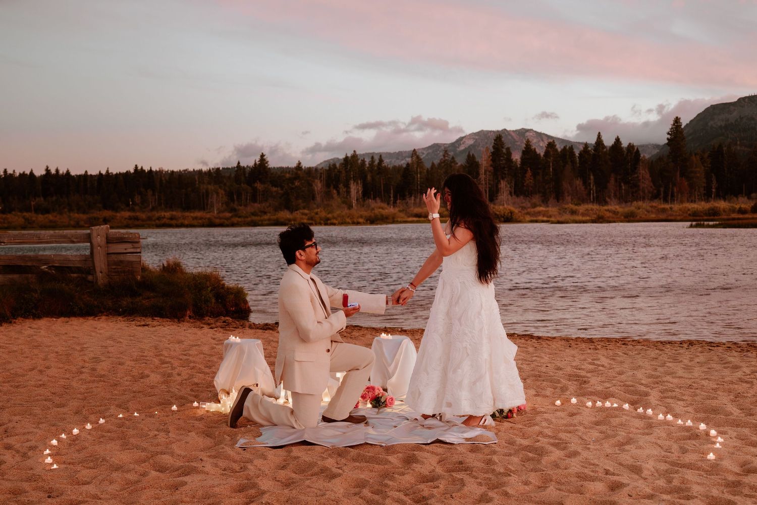 Man proposing to woman on sandy beach at sunset with candles, mountains and lake in background.