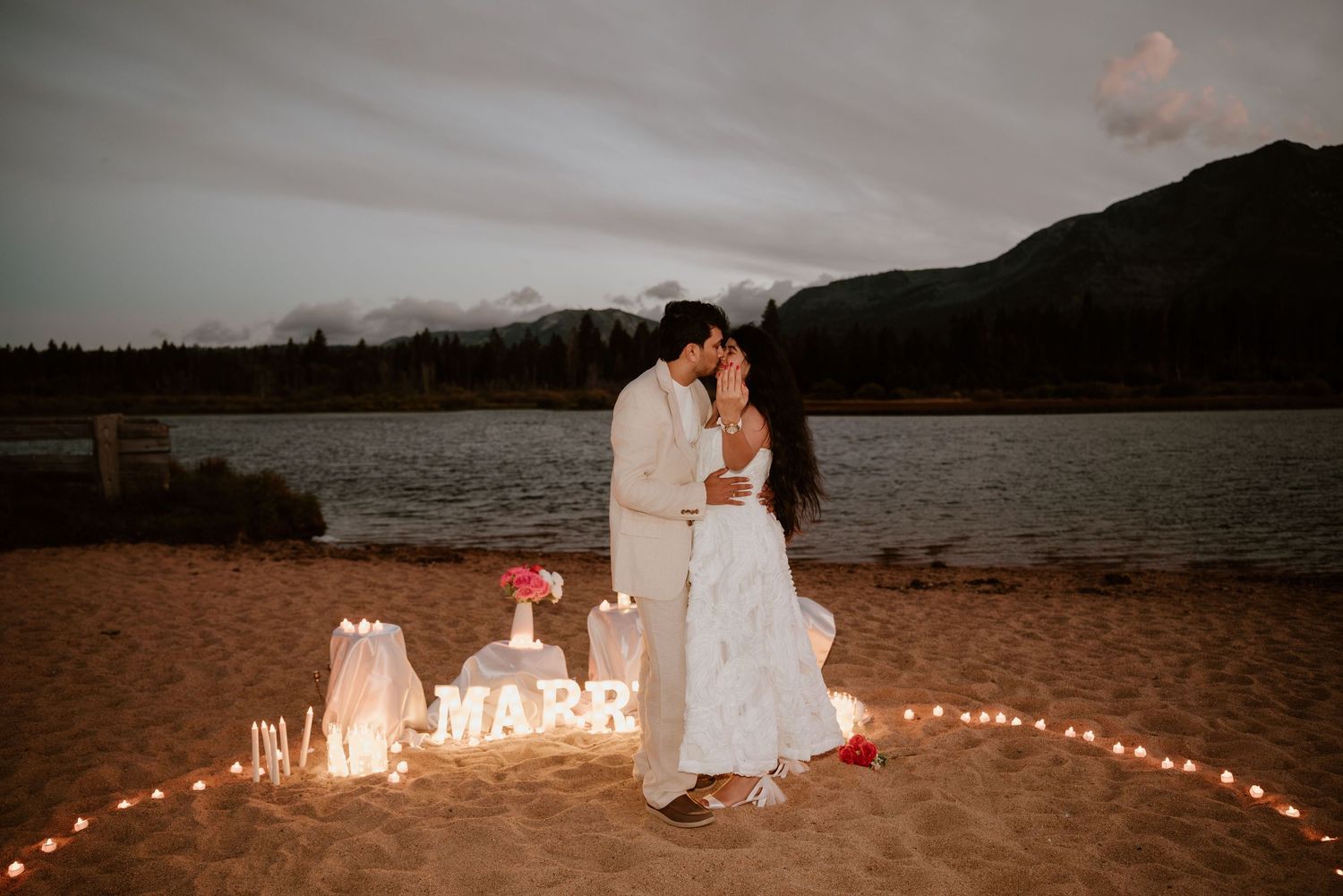 Couple kissing on sandy beach at dusk with glowing MARRY letters and candles by a lake and mountains.