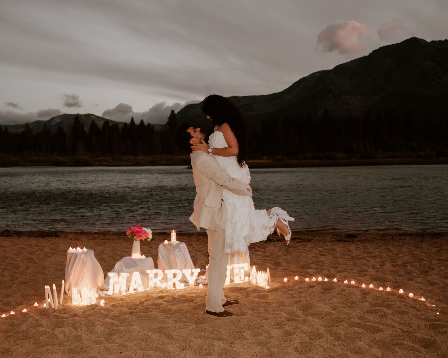 Couple sharing a romantic beach proposal at dusk with lit candles and MARRY ME sign by a mountain lake.