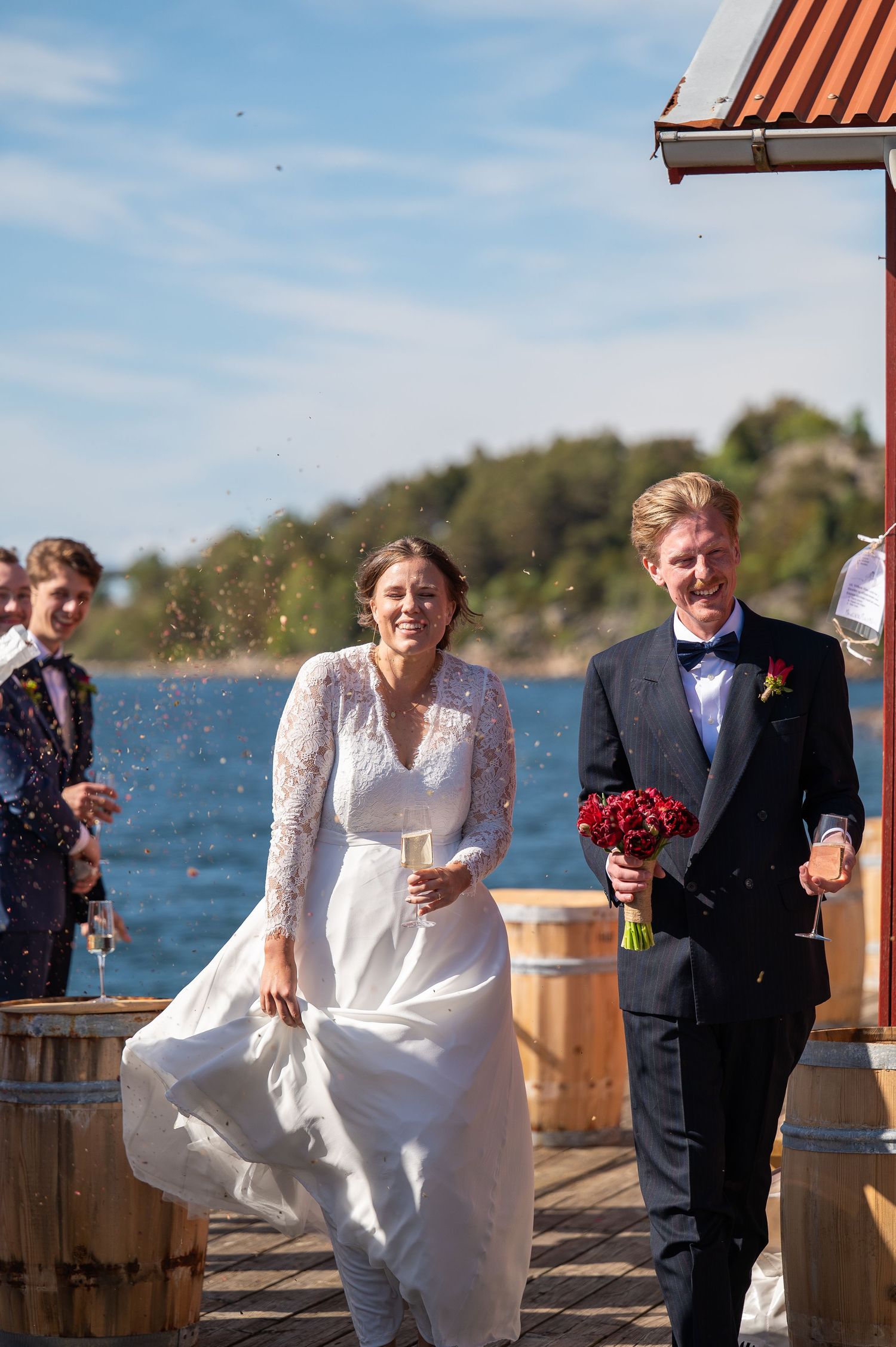 Bride and groom walk on a waterfront dock, smiling, as guests toss confetti on a sunny wedding day.
