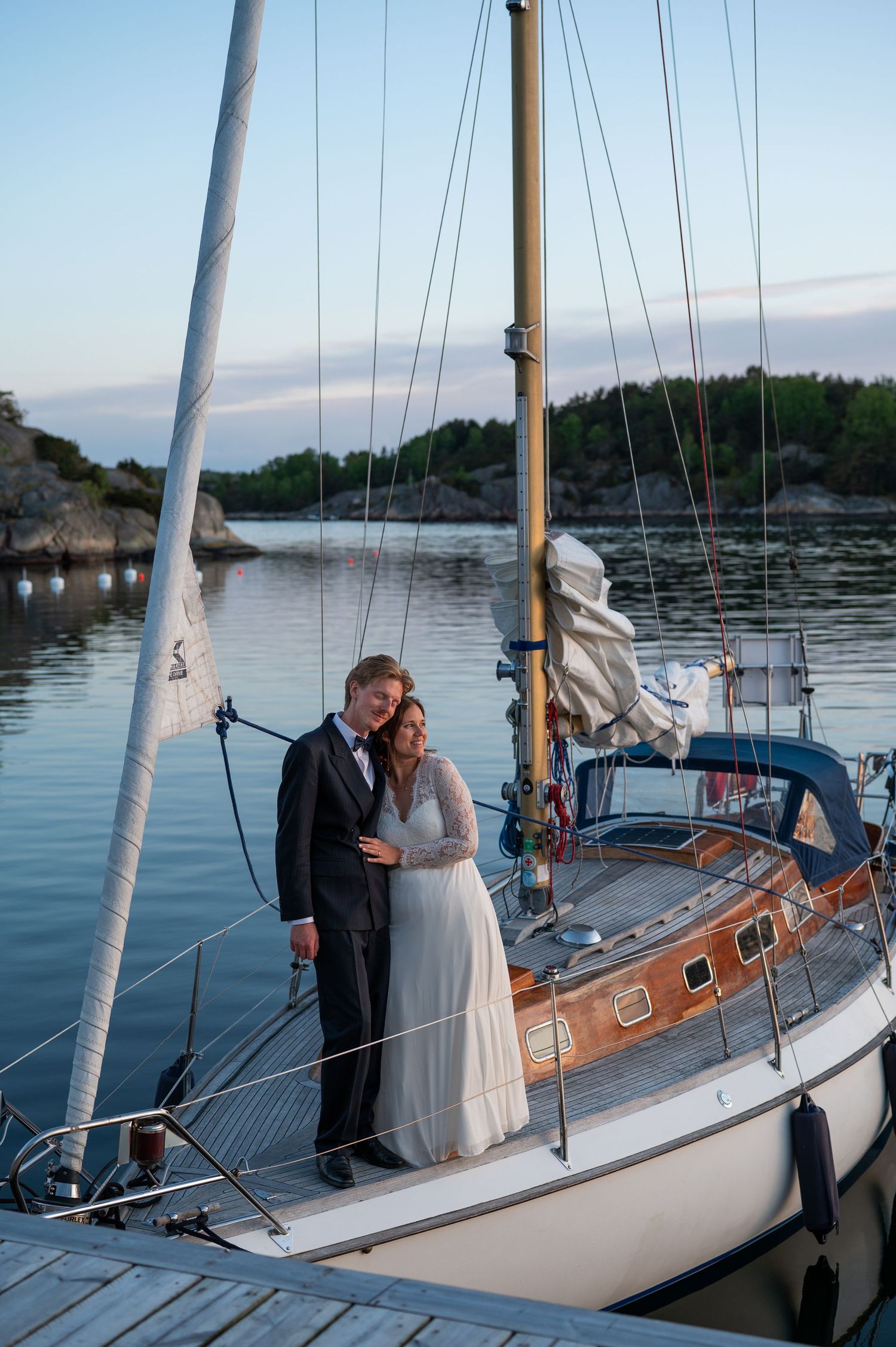 Bride and groom embracing on a classic sailboat at dusk, with calm waters and rocky islands in the background.
