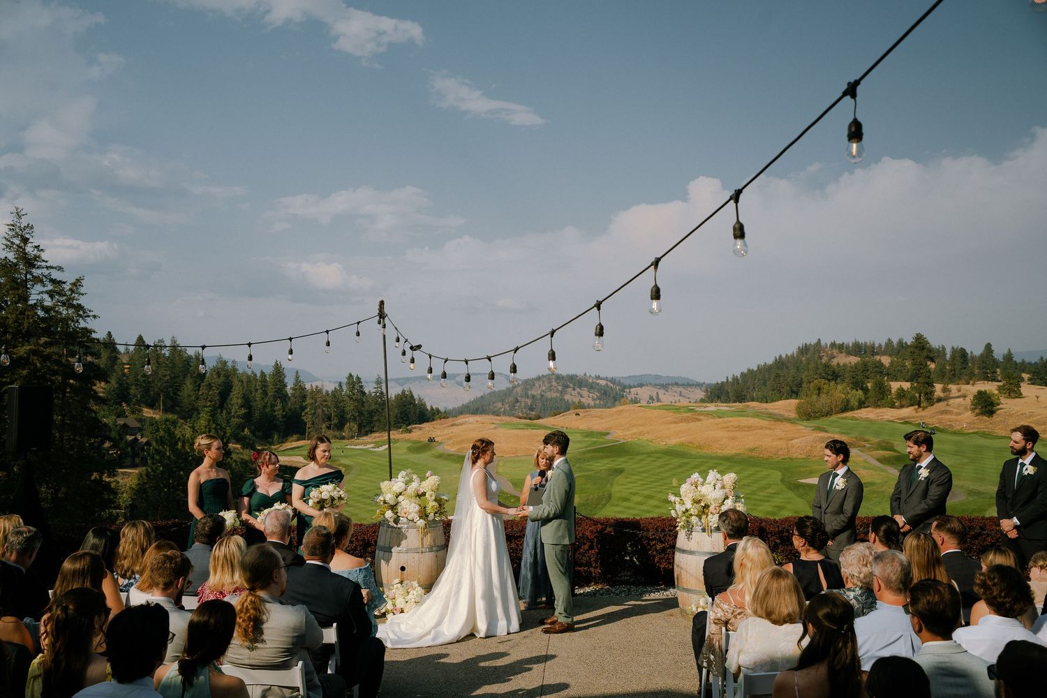 Outdoor wedding ceremony on a terrace overlooking a scenic golf course, with string lights and guests seated under a blue sky.