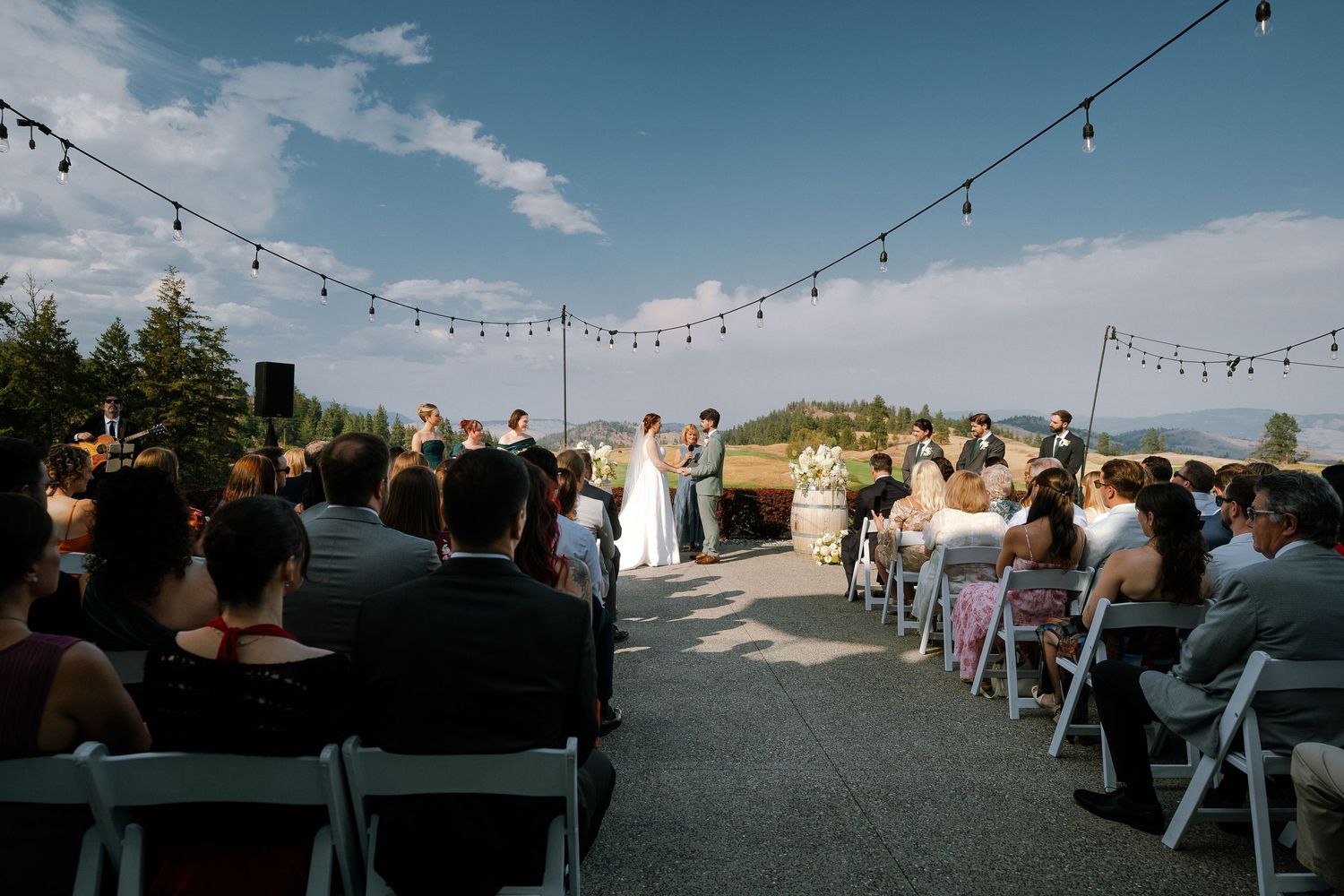 Outdoor wedding ceremony with string lights, guests seated in white chairs, couple at altar with mountain views.