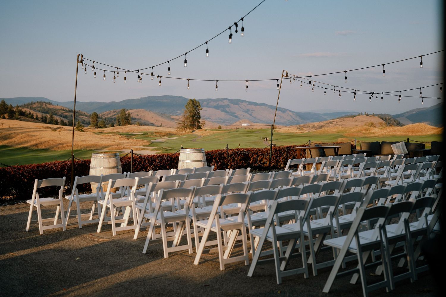 Outdoor wedding ceremony setup with white folding chairs, wine barrel decor, string lights, and scenic valley views.
