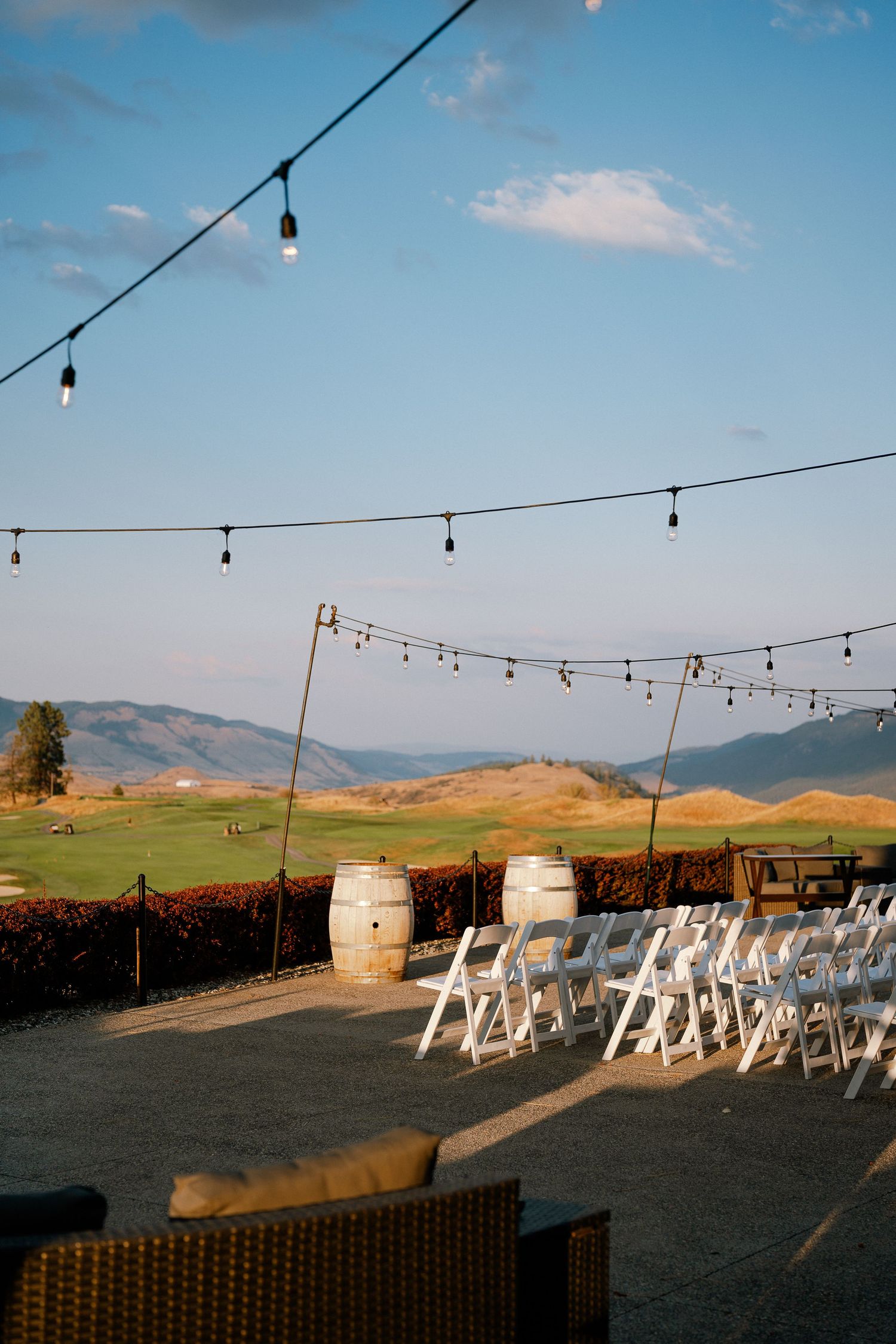 Outdoor wedding venue with white chairs, wine barrels, string lights, and scenic mountain views under a clear blue sky.