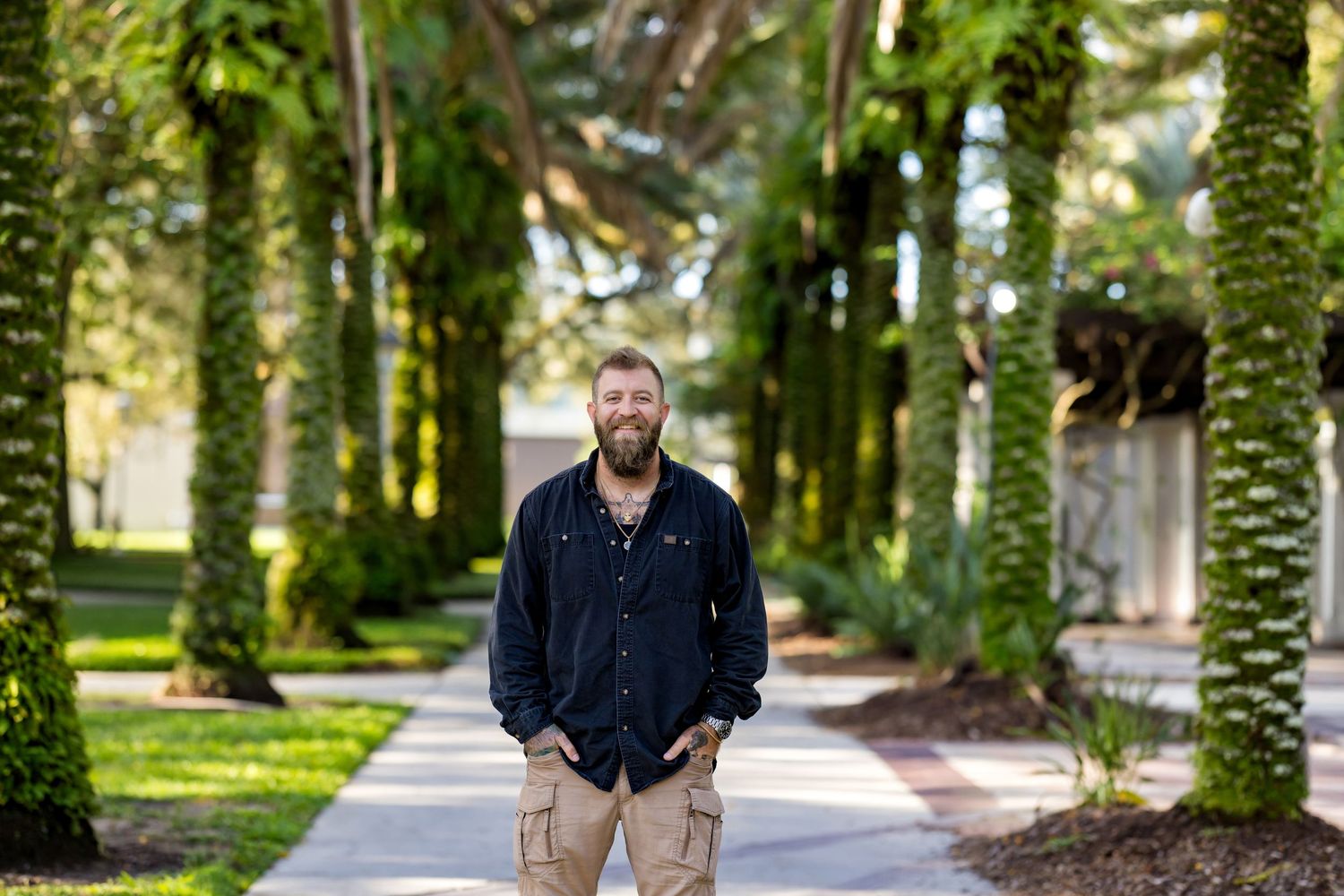 Bearded man in black shirt and khaki pants stands on a palm tree-lined pathway in a tropical setting.