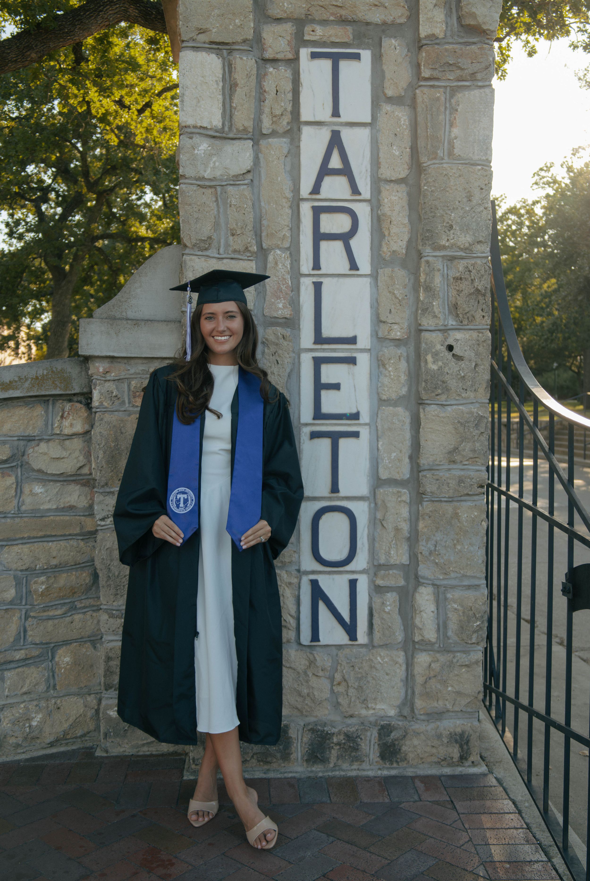 Graduate in cap and gown poses proudly beside Tarleton State University stone entrance sign.
