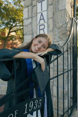Smiling graduate in black gown and blue stole leans on iron gate with 1935 inscription at stone university entrance.
