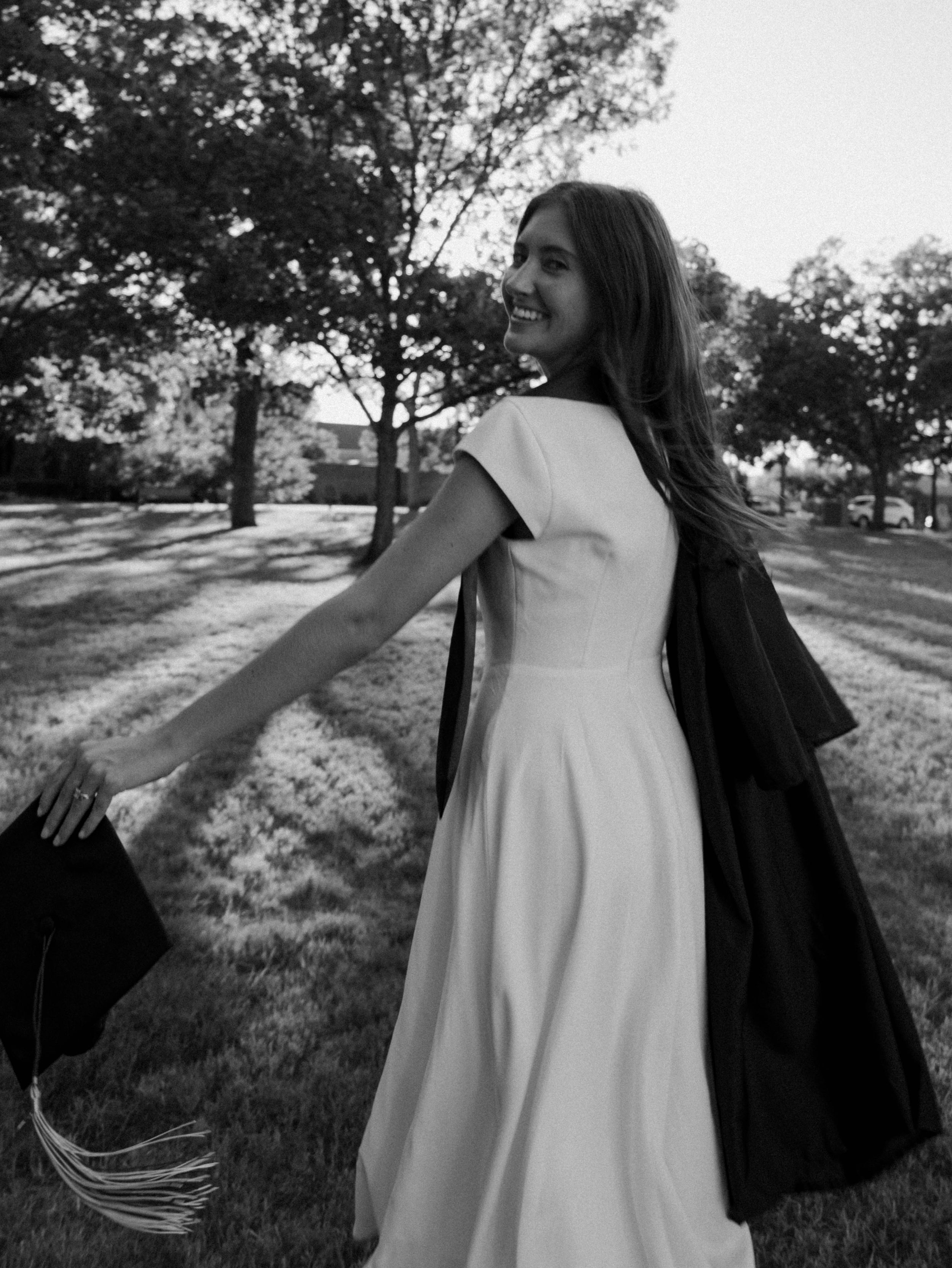 Black and white photo of a smiling graduate in white dress holding cap and gown outdoors on a sunny day.