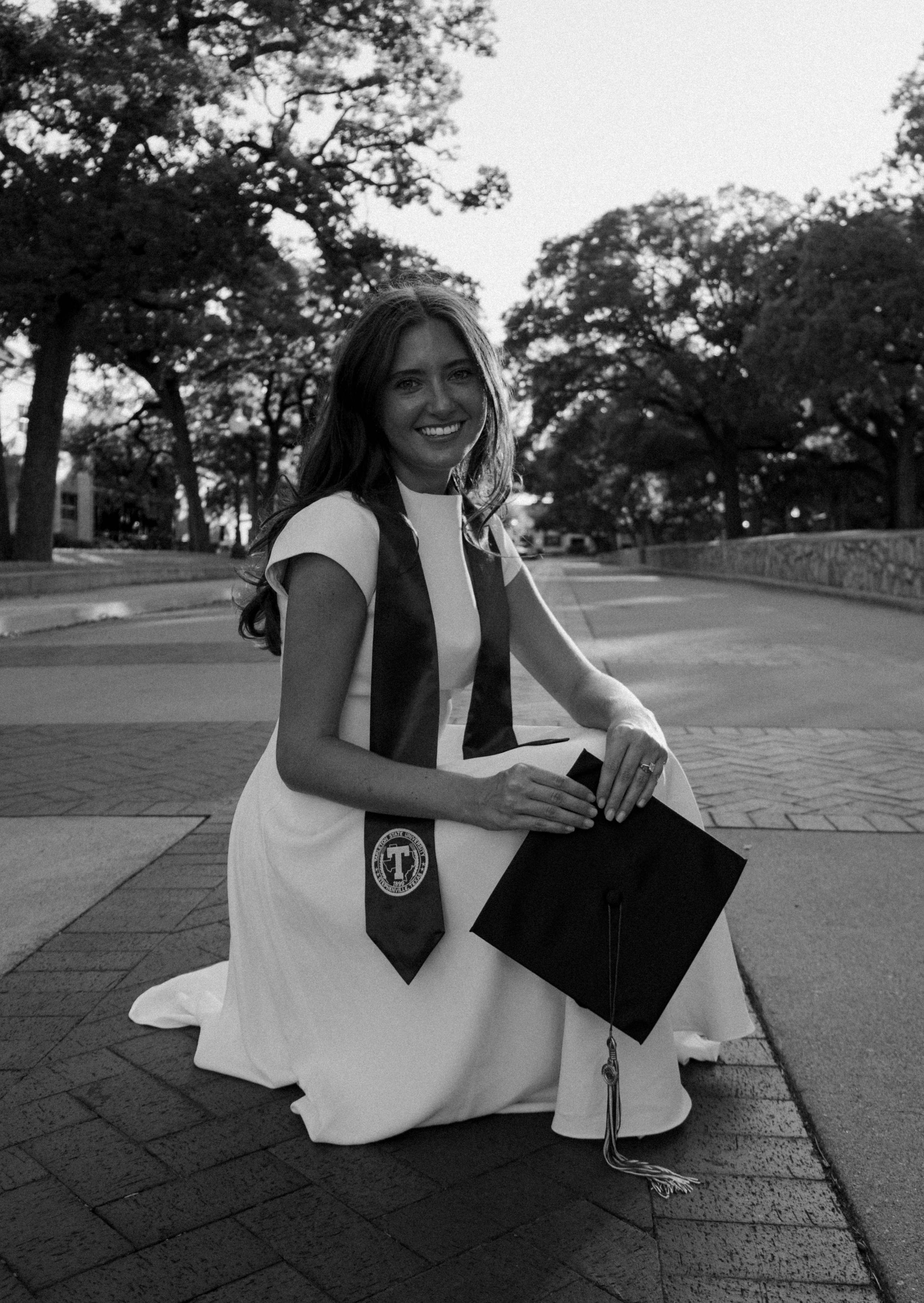 A smiling young woman in a white dress and graduation stole holds her cap while kneeling on a brick pathway outdoors.