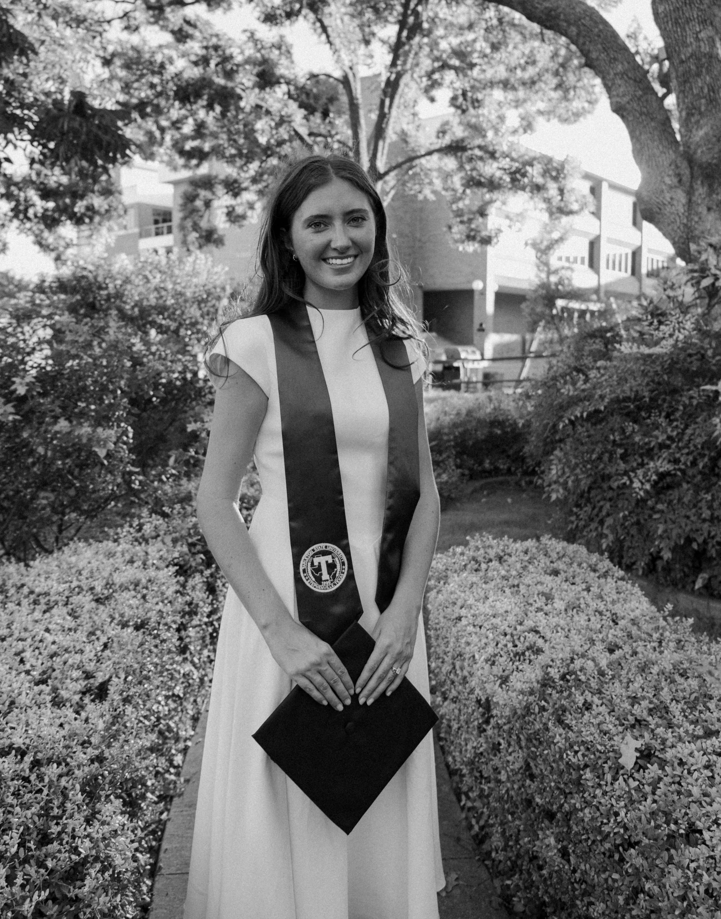 Young woman in white graduation dress and black academic stole smiles outdoors among lush greenery.